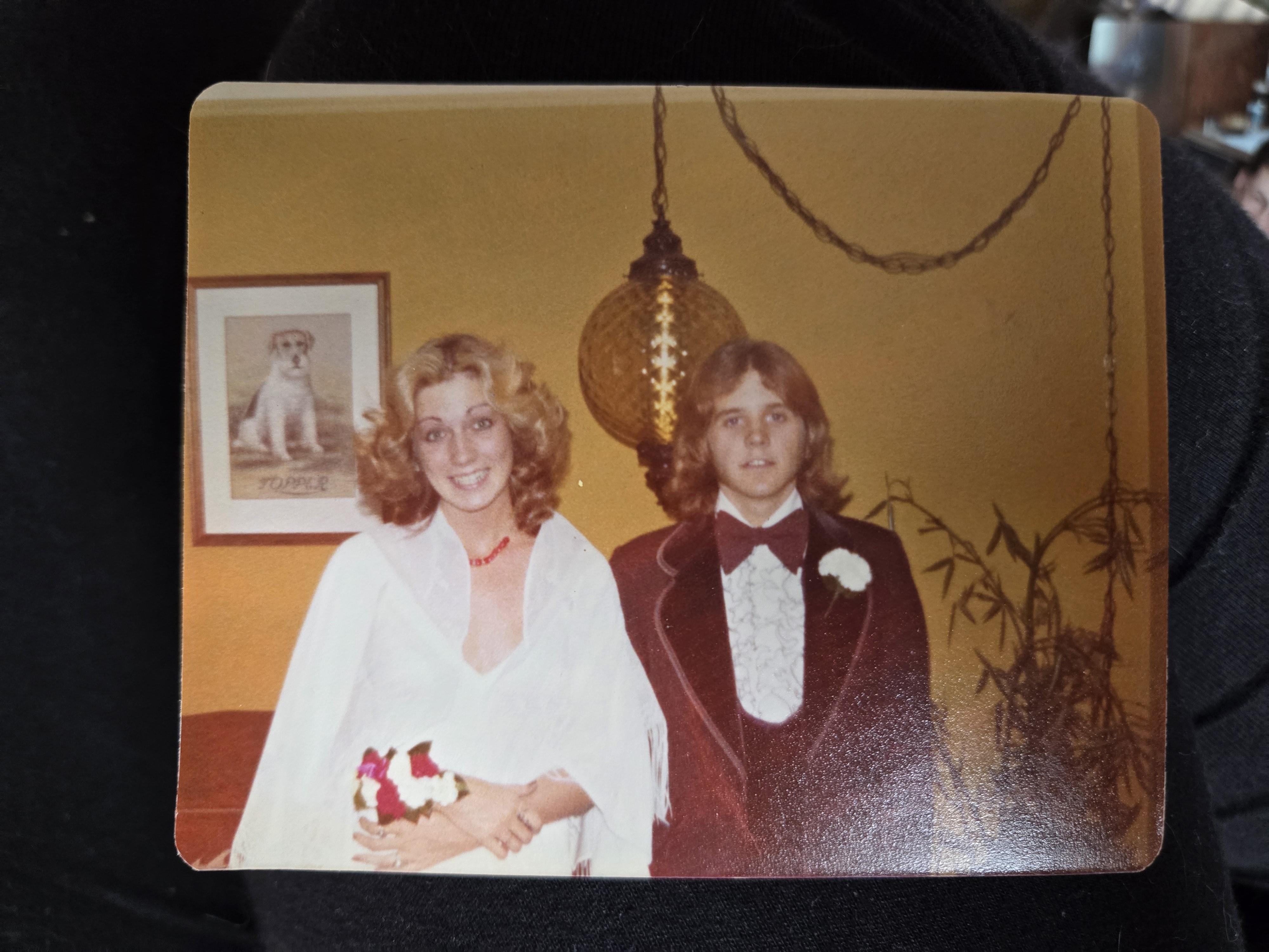 A couple in 1970s formal attire poses elegantly in a warmly lit room.