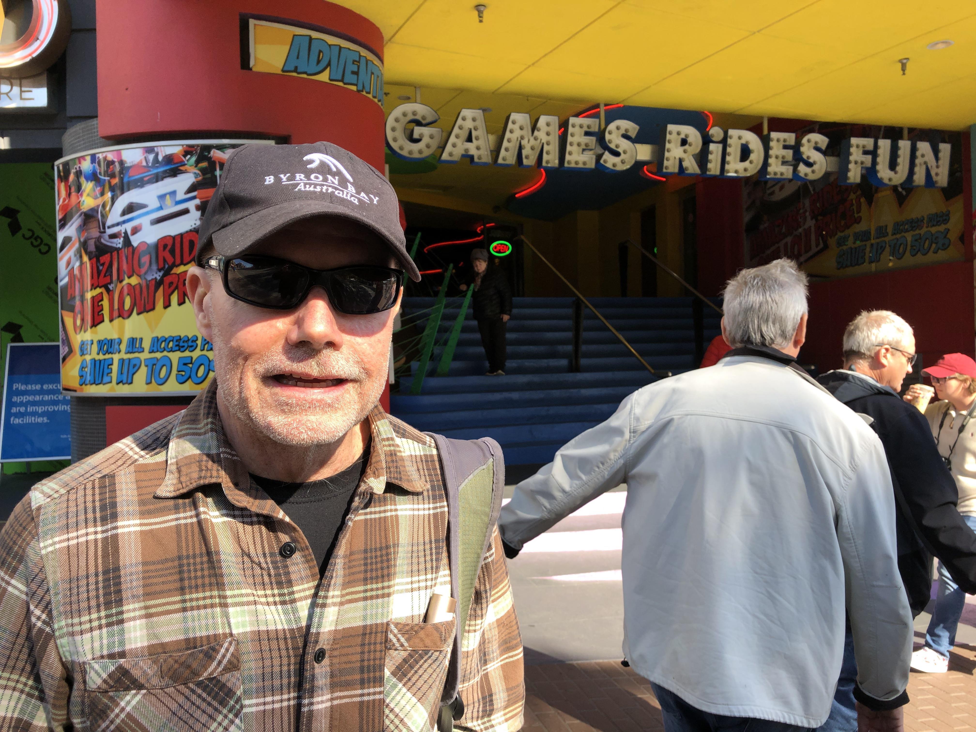 A man wearing sunglasses and a plaid shirt stands near a vibrant amusement center entrance.