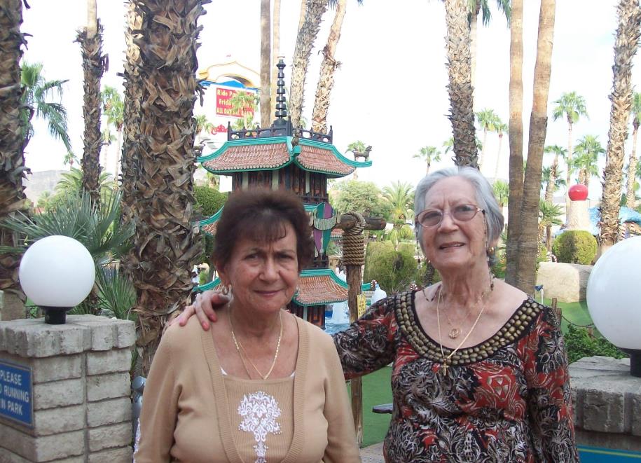 Elderly women smile at the camera while standing in a vibrant garden with palm trees.
