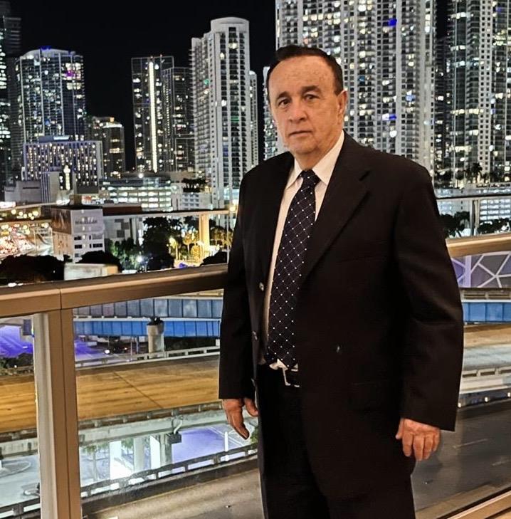 A distinguished man poses near a modern railing with Miami's sparkling skyline behind him at night.