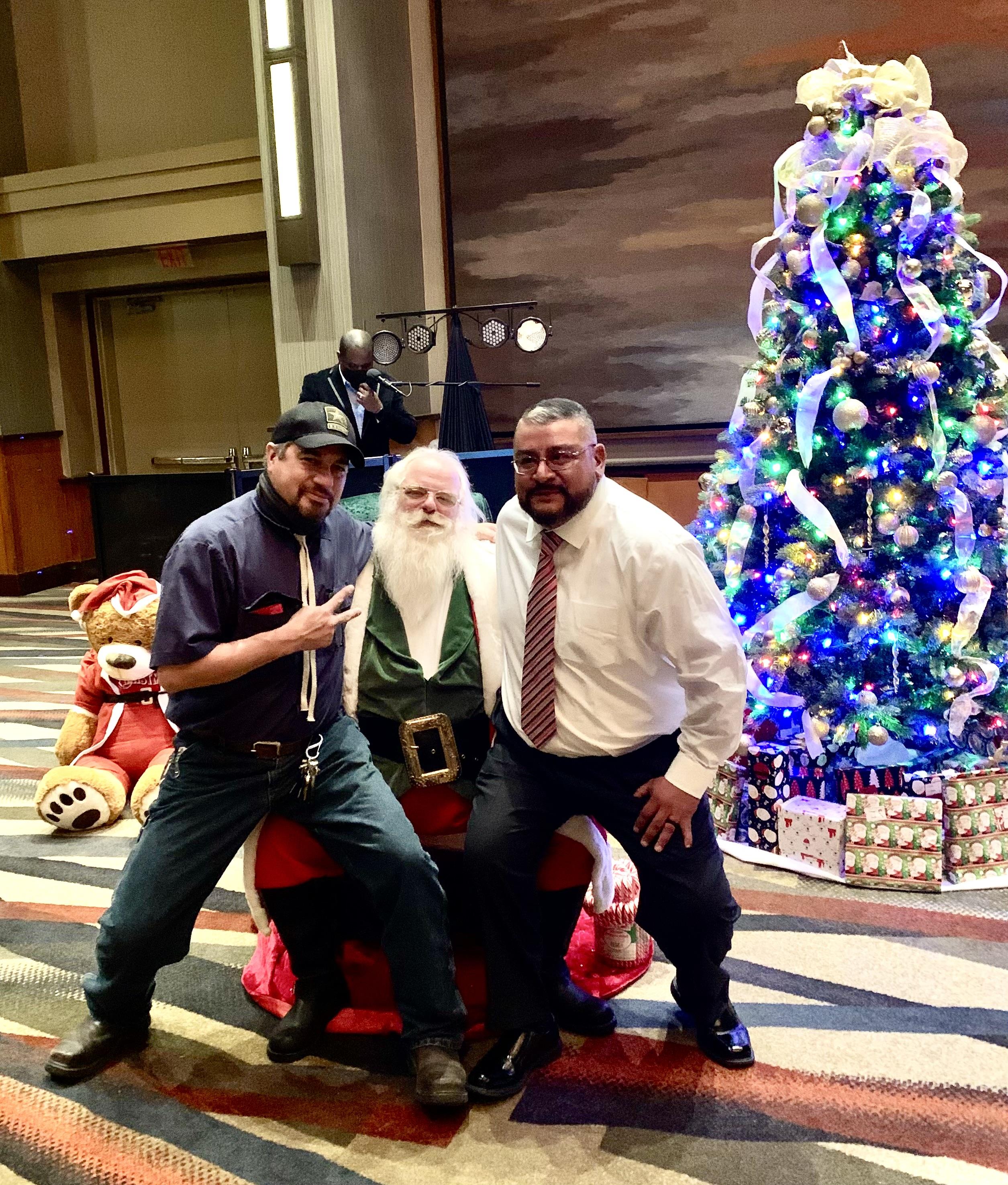 Two friends joyfully pose with Santa Claus in front of a decorated Christmas tree and gifts.