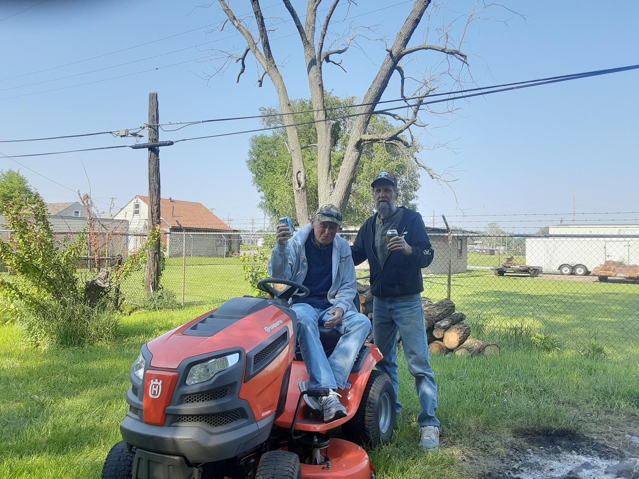 Two men stand beside a lawn mower, smiling and enjoying the sunny afternoon in a yard.