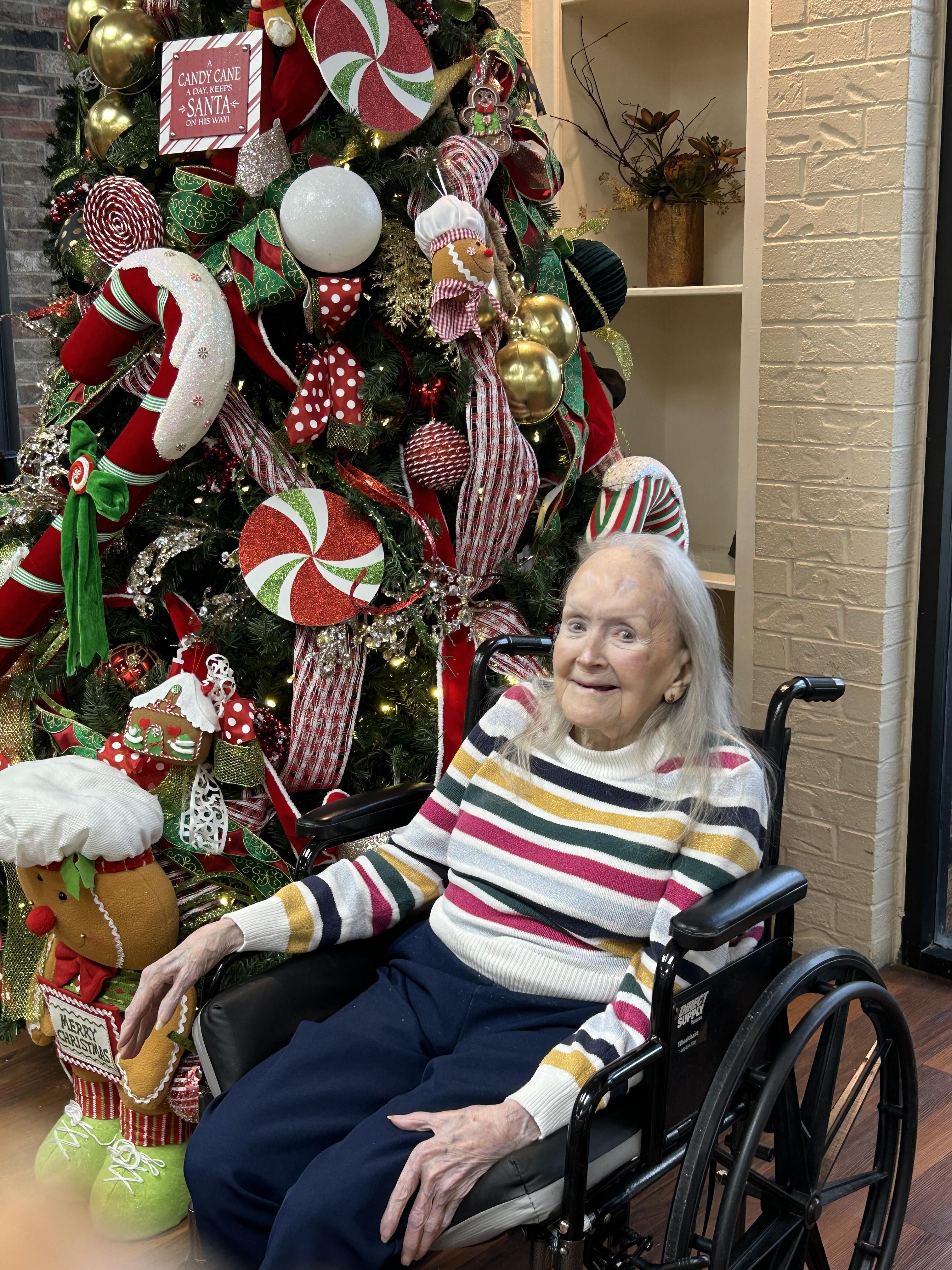 A joyful elderly woman in a wheelchair smiles next to a brightly decorated Christmas tree.