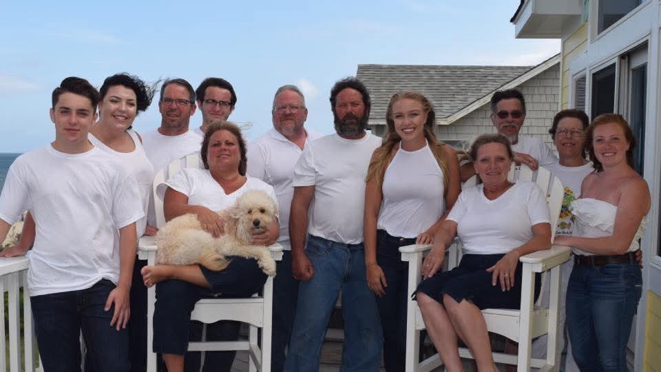 A large group of people, wearing white shirts, smiles together on a balcony enjoying summer.