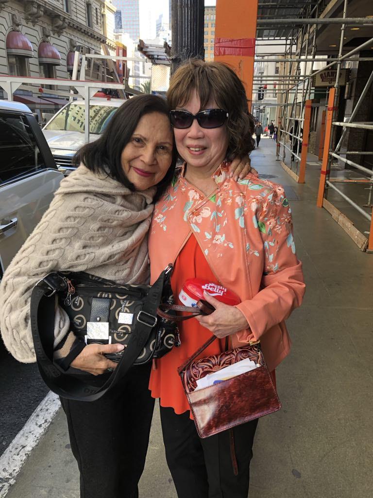 Two women smile and embrace on a busy city street while carrying fashionable handbags.