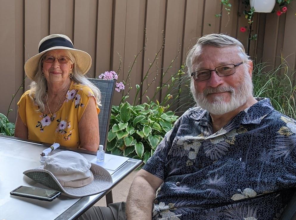 Two friends share laughs at a garden table surrounded by greenery on a sunny afternoon.