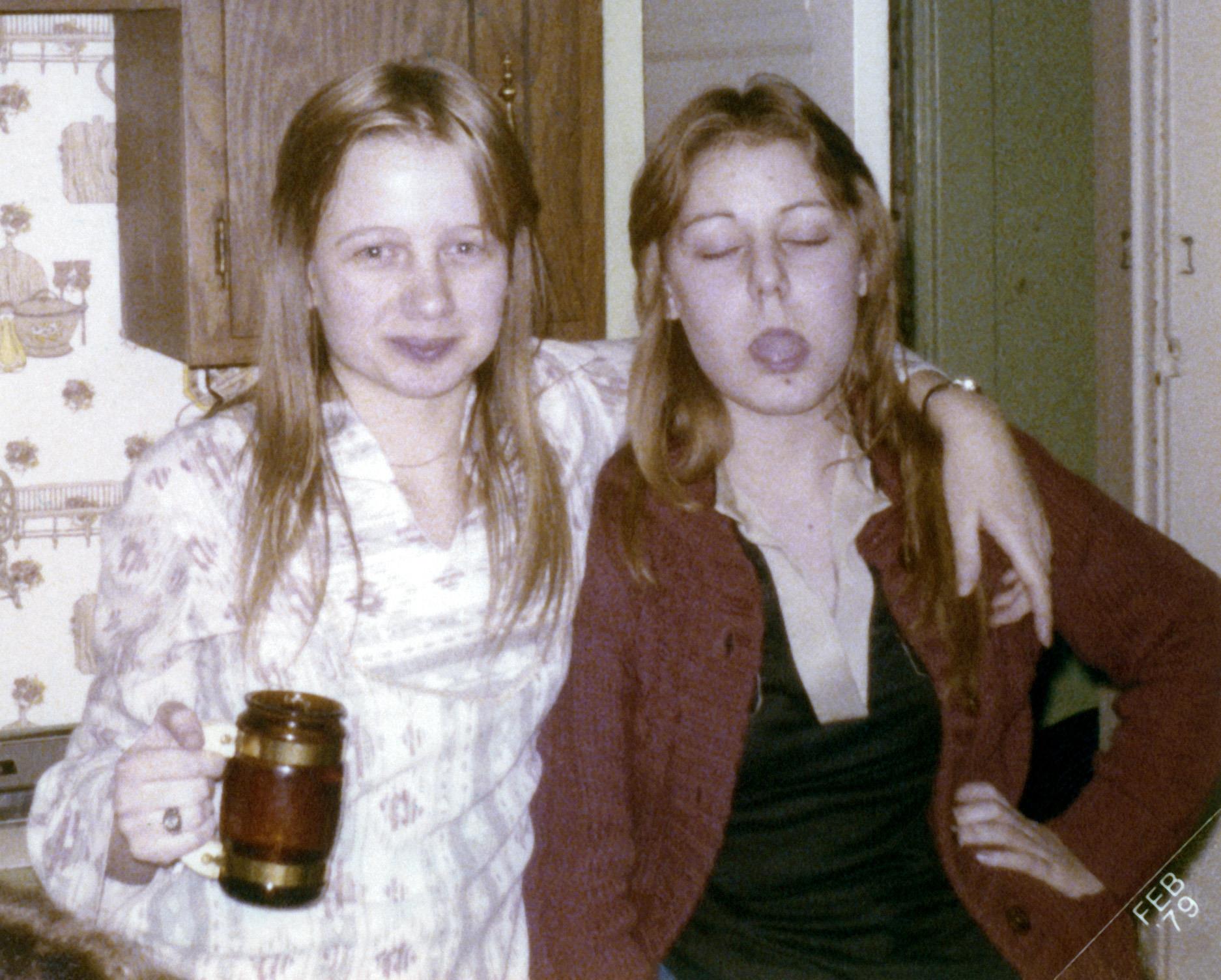 Two girls stand close together, one holding a drink while both smile playfully indoors.