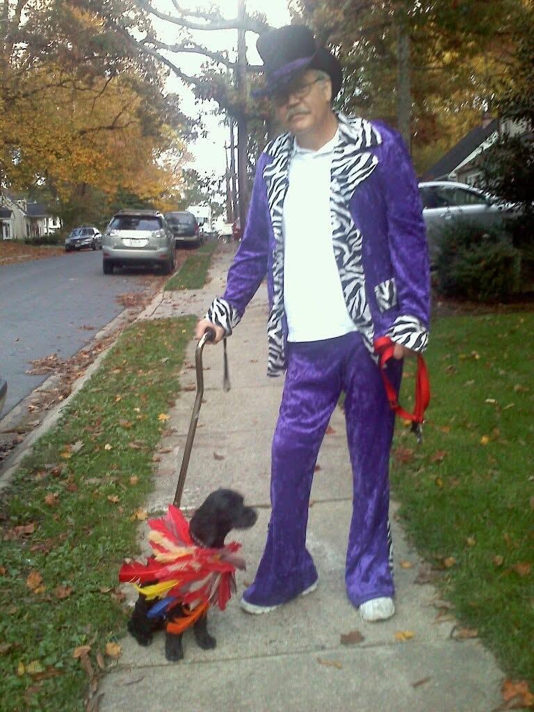 A man in a purple suit walks his dog in a bright costume on a brisk autumn day.