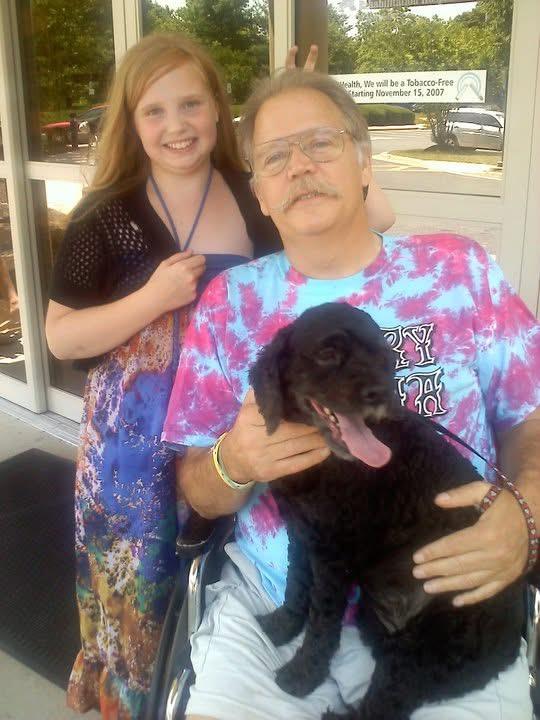 A man in a wheelchair enjoys a sunny day with a smiling girl and a playful black dog.