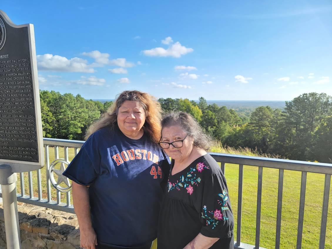 Two women stand closely together, smiling at a scenic viewpoint with expansive greenery.