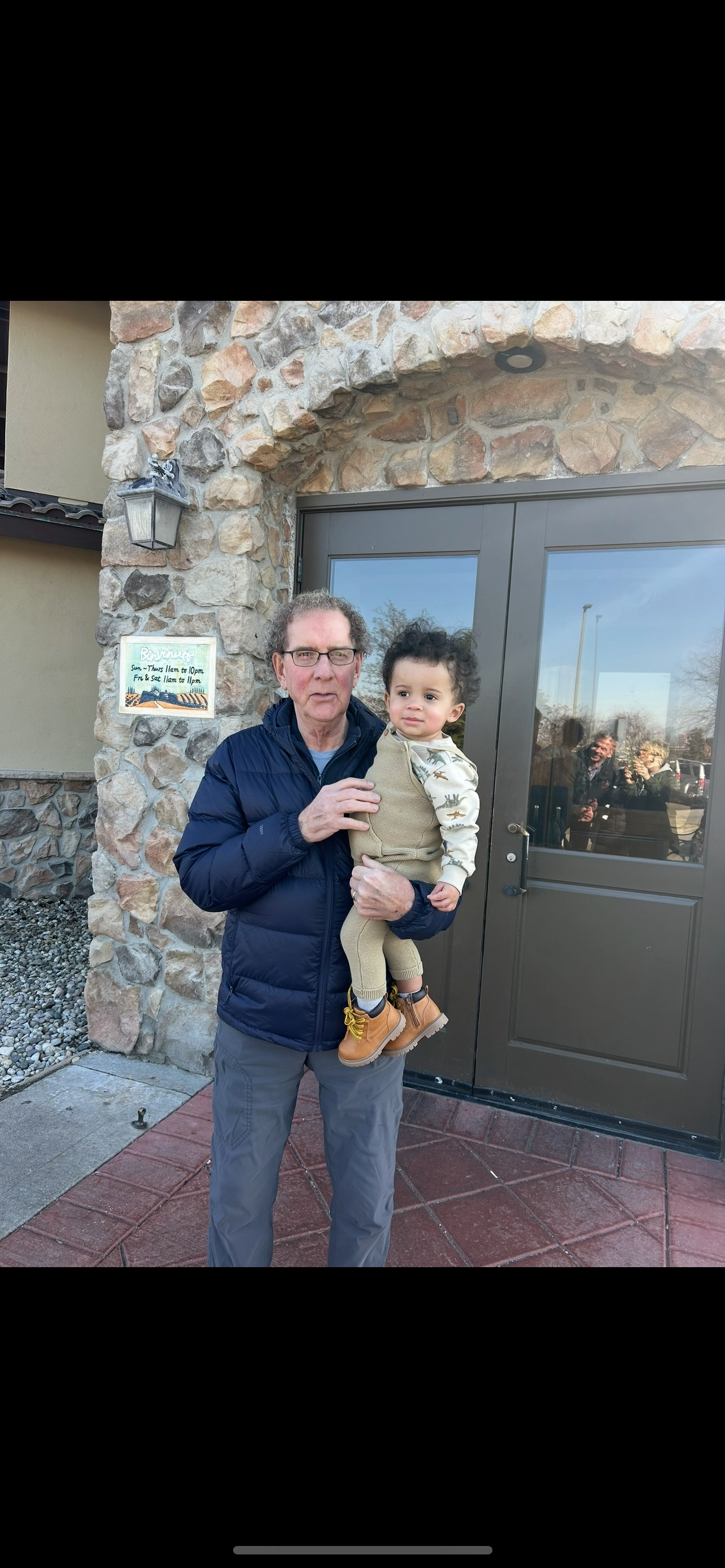 A delighted grandfather embraces his young grandchild in front of a charming stone structure.