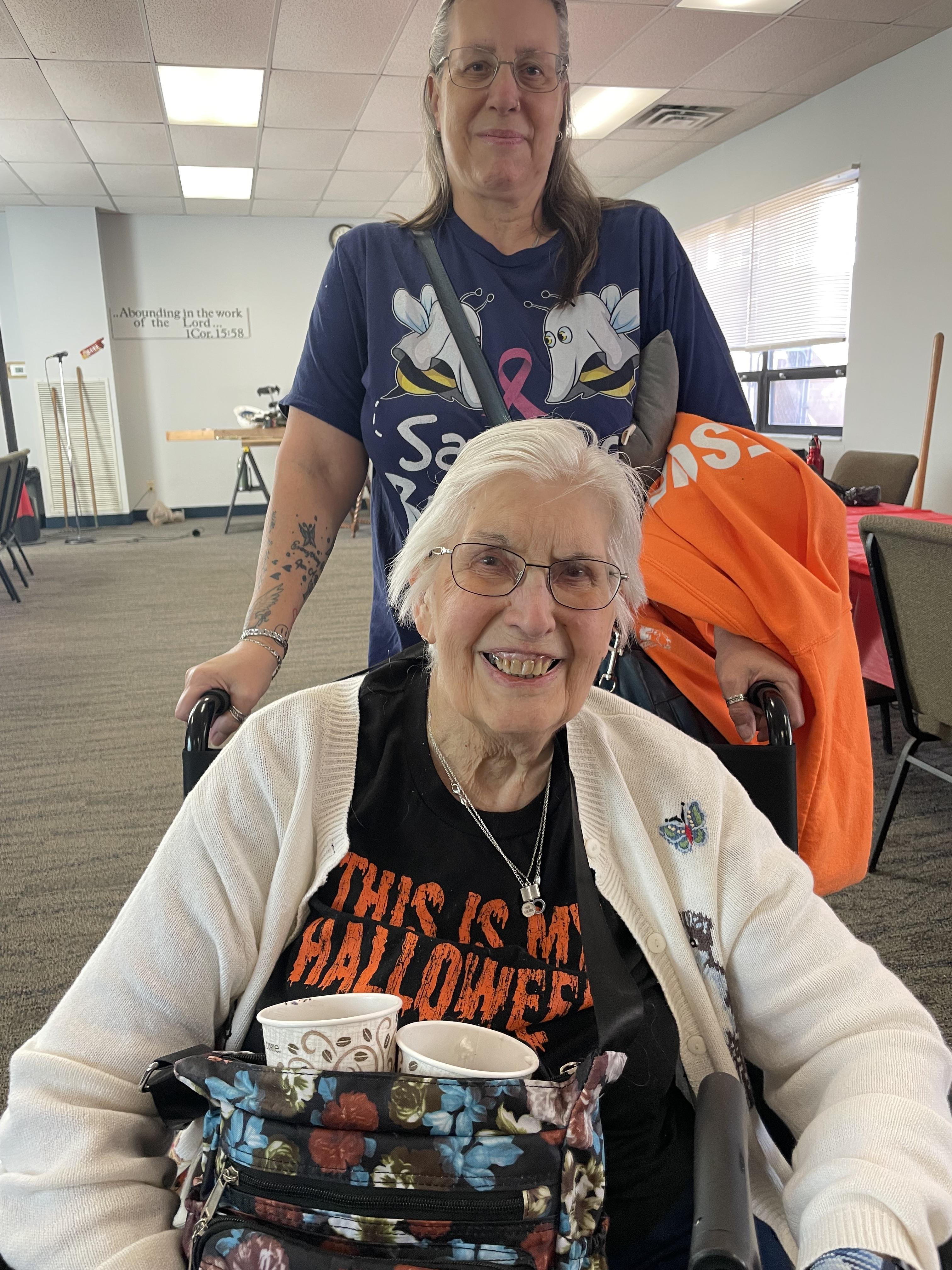 Senior woman in a wheelchair smiles with her caregiver at a Halloween community event.