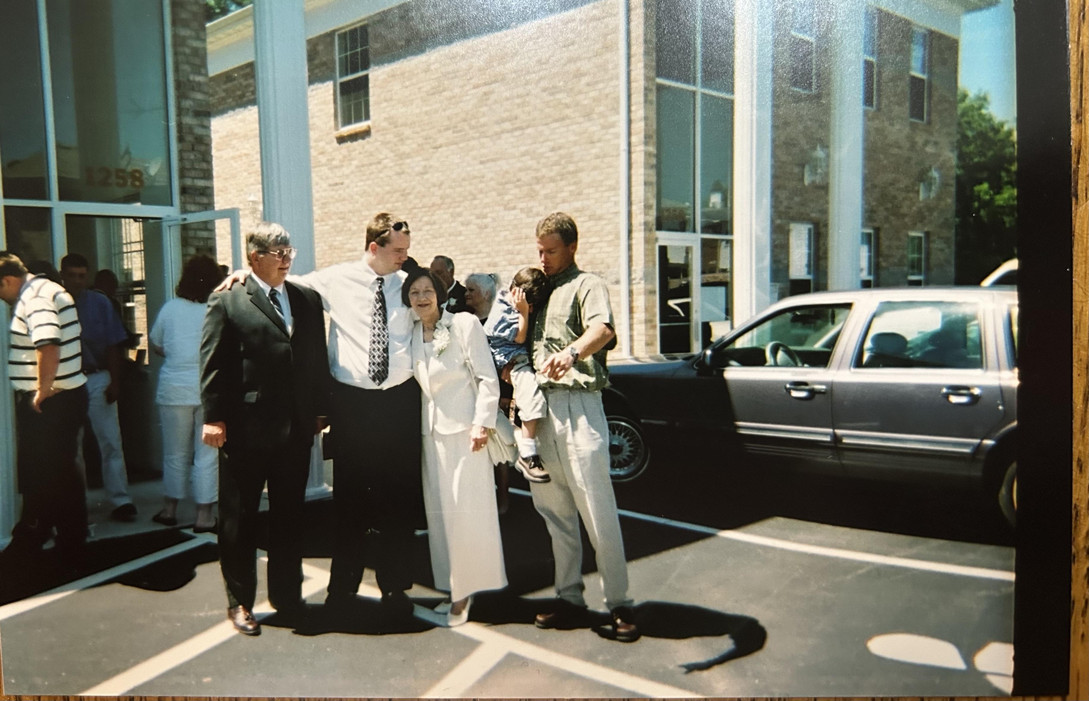 Four adults stand together, smiling outside a building while others mingle in the background.