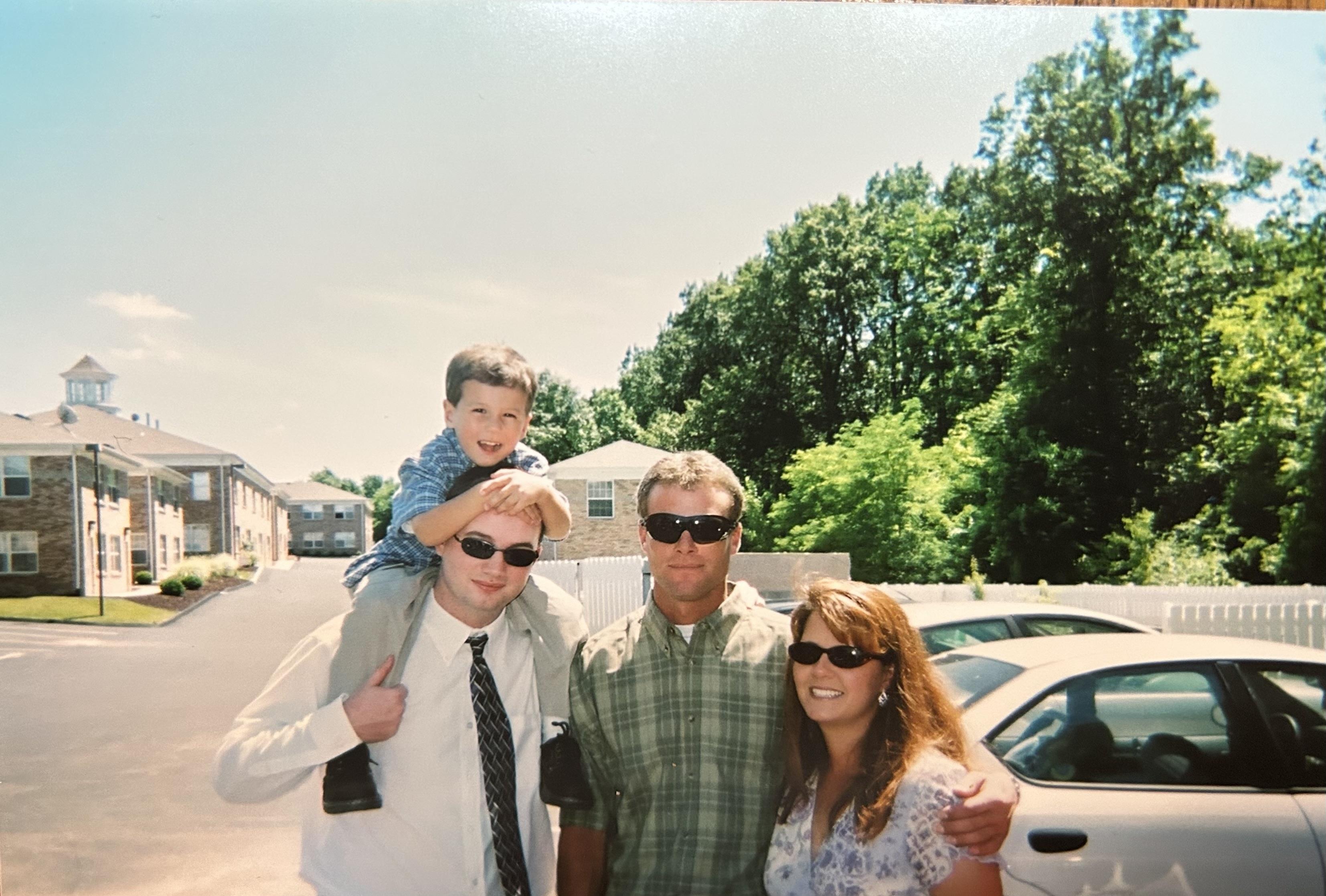 Four people pose for a cheerful group photo outside an apartment complex under bright sunshine.