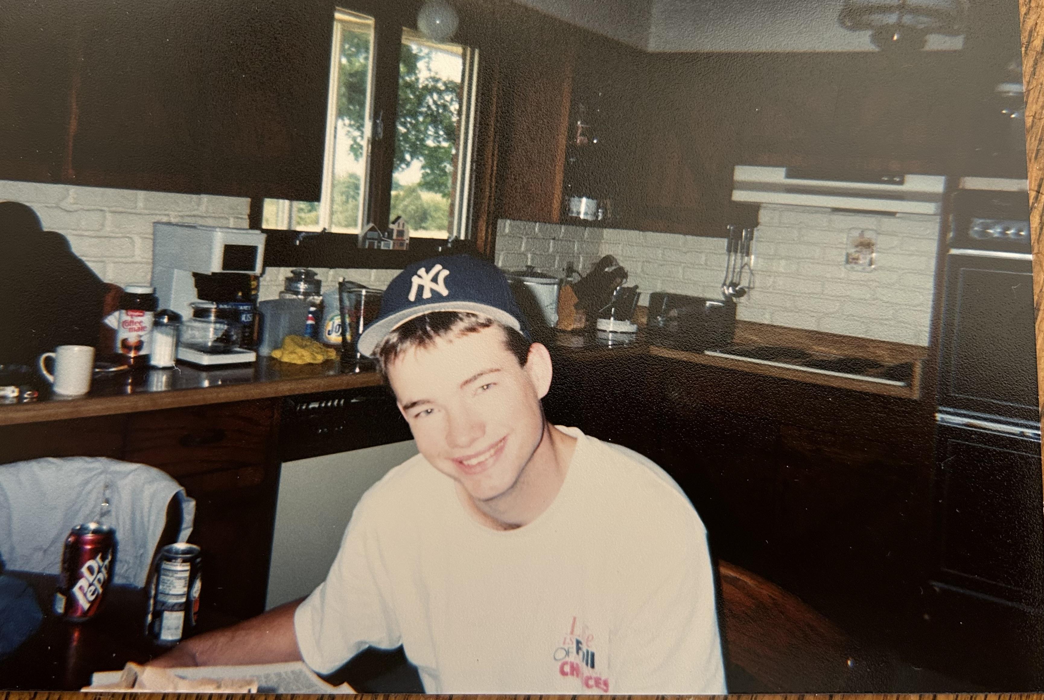A young man sits at a kitchen table, smiling while working on a project. Sunlight fills the room.