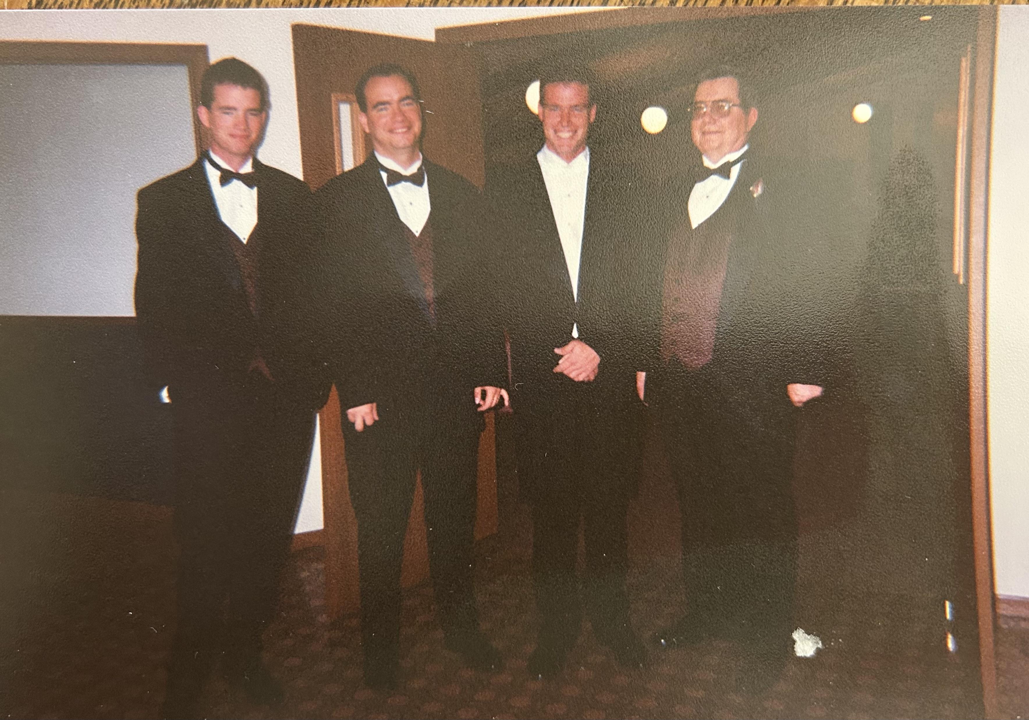 Four men in tuxedos stand together near an entrance, smiling and enjoying a special occasion.