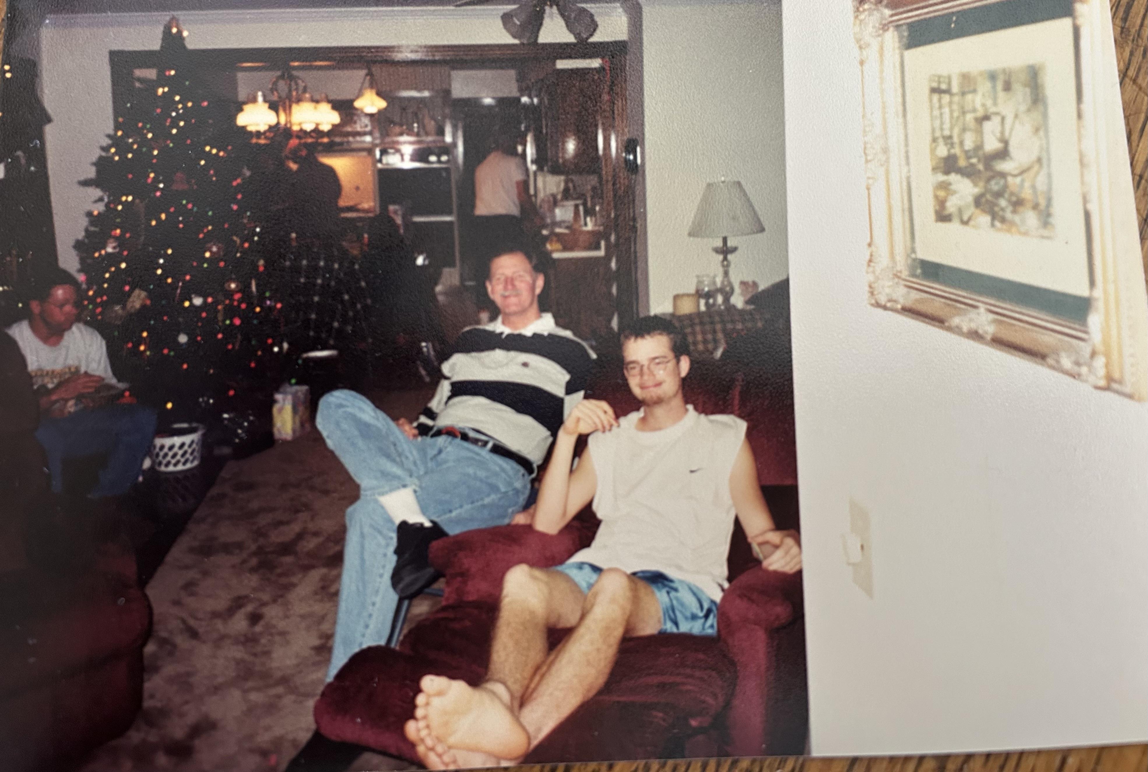 Two men relax on a couch while a Christmas tree sparkles in the background.