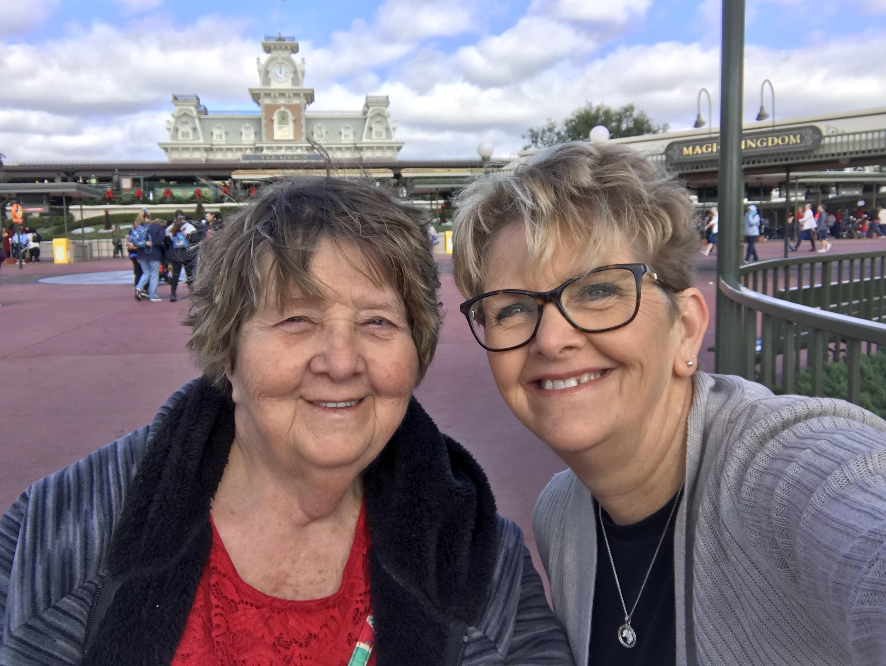 Two women smile together at the entrance of a popular amusement park on a clear day.