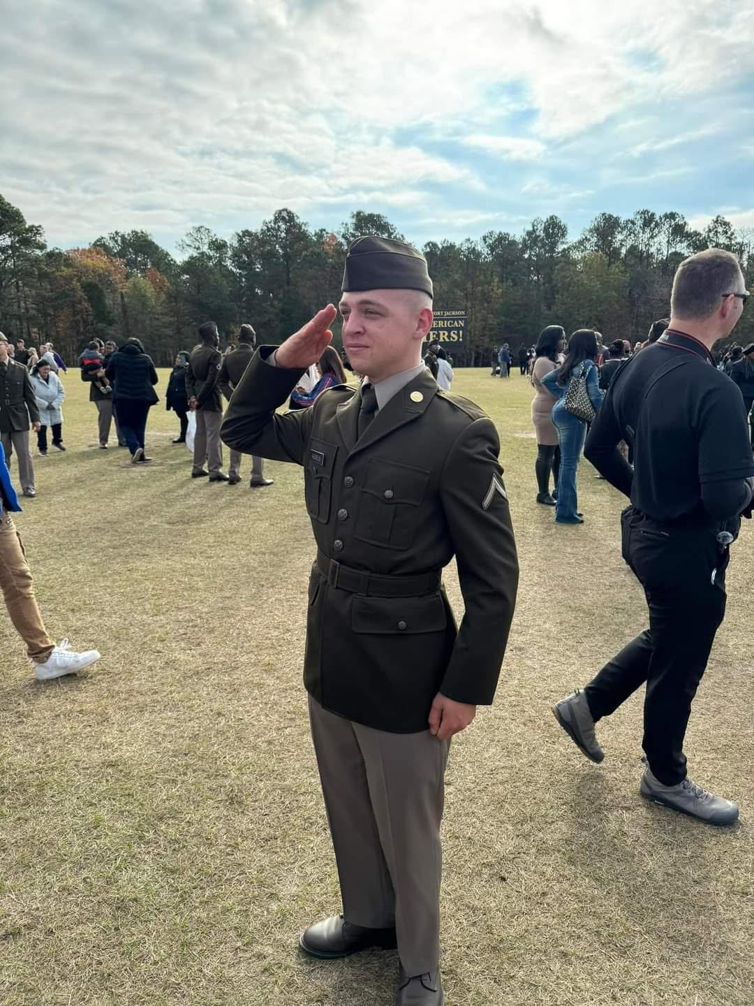 A young soldier in uniform salutes while surrounded by a gathering of people outdoors.
