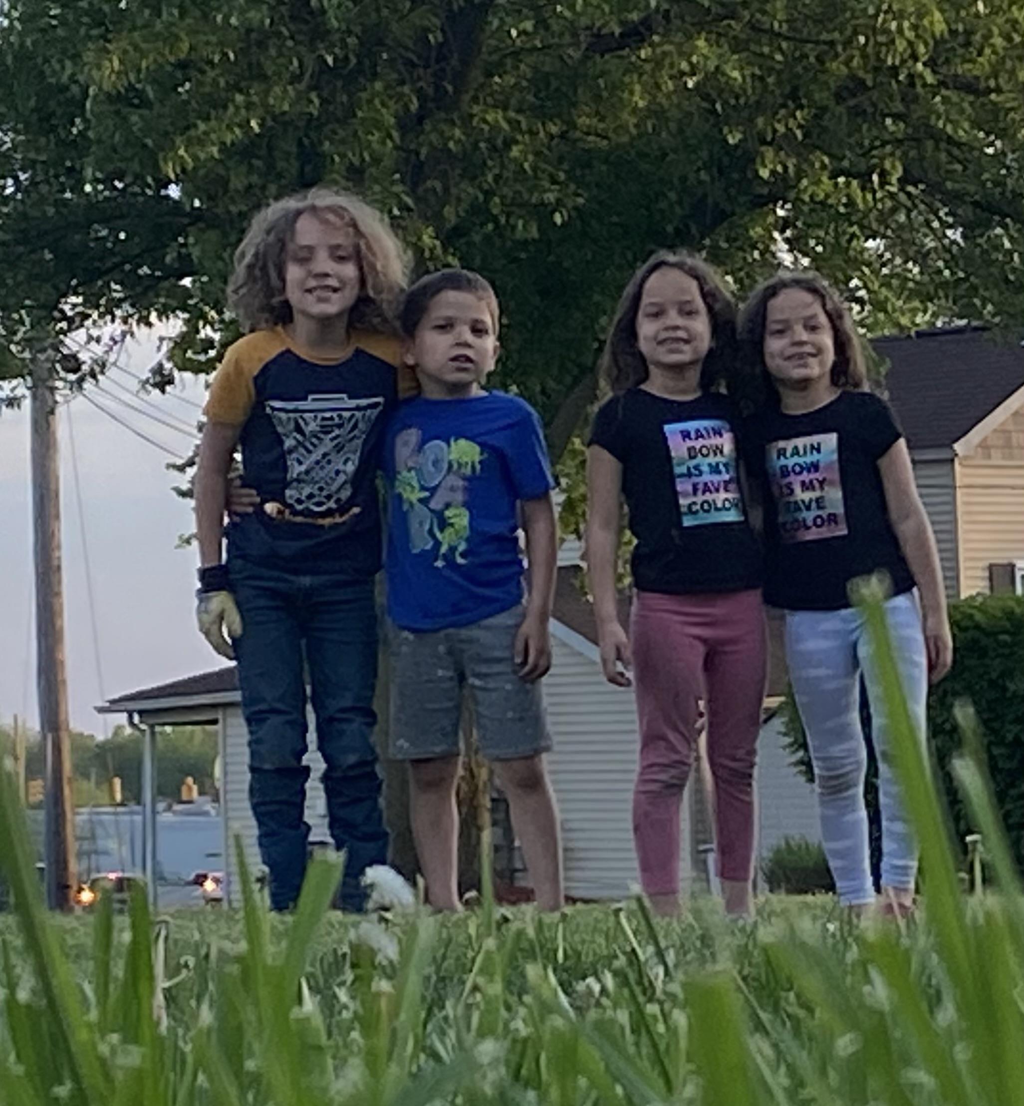 Four joyful children posing together in a grassy area near homes during the evening.