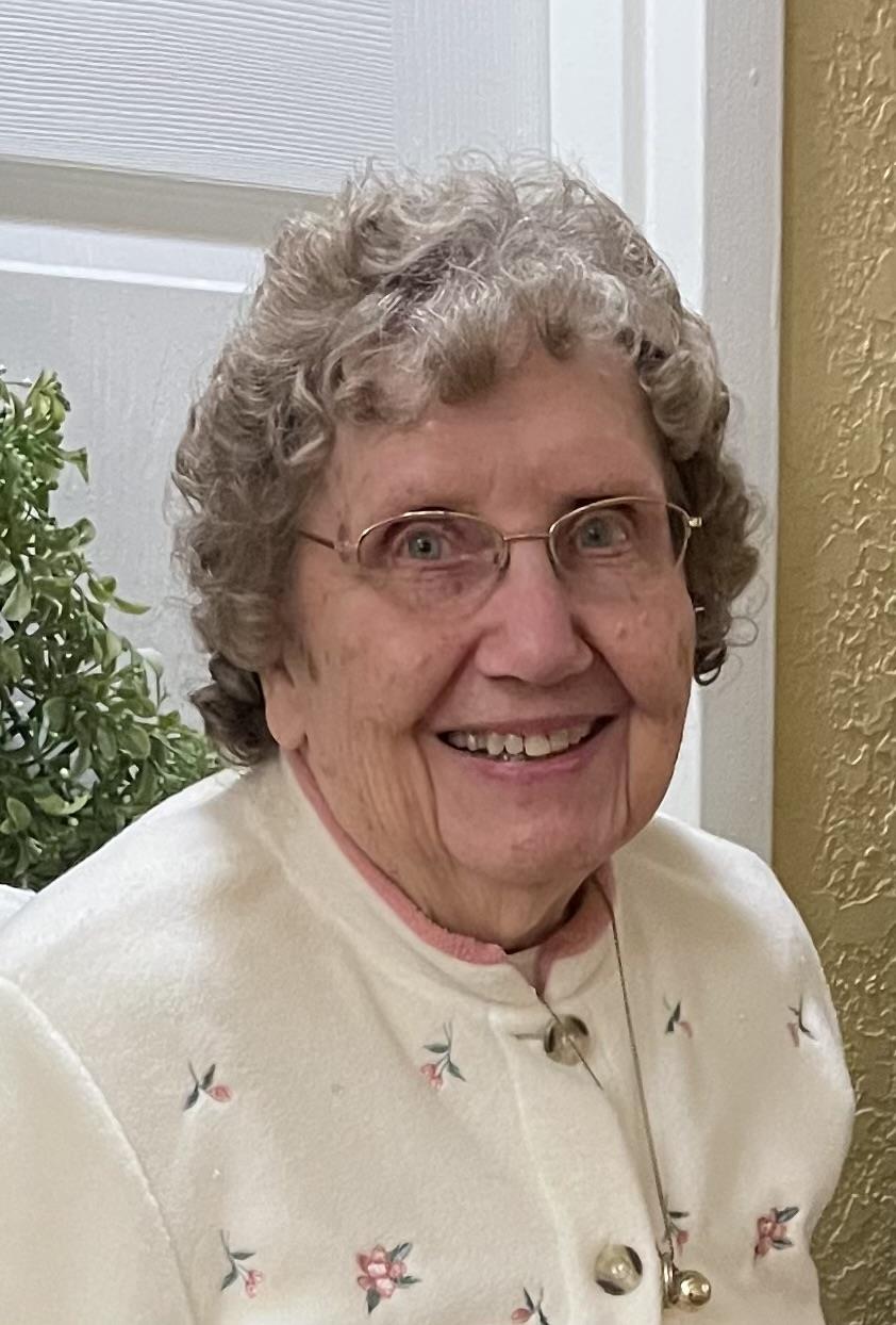 Elderly woman with glasses smiles warmly, seated indoors amid soft lighting and plants.