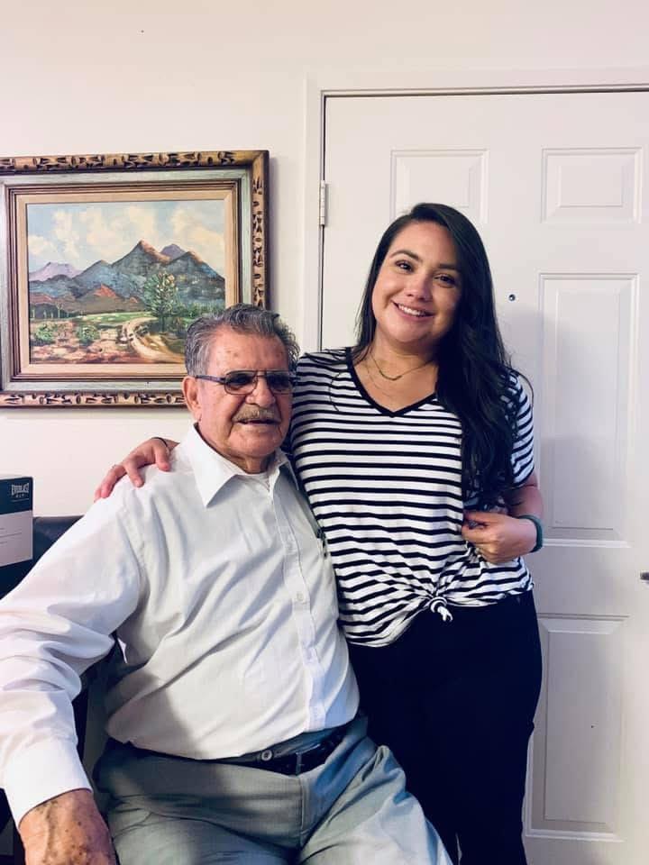 A senior man sits comfortably, smiling beside a young woman in a casual outfit, conveying warmth.