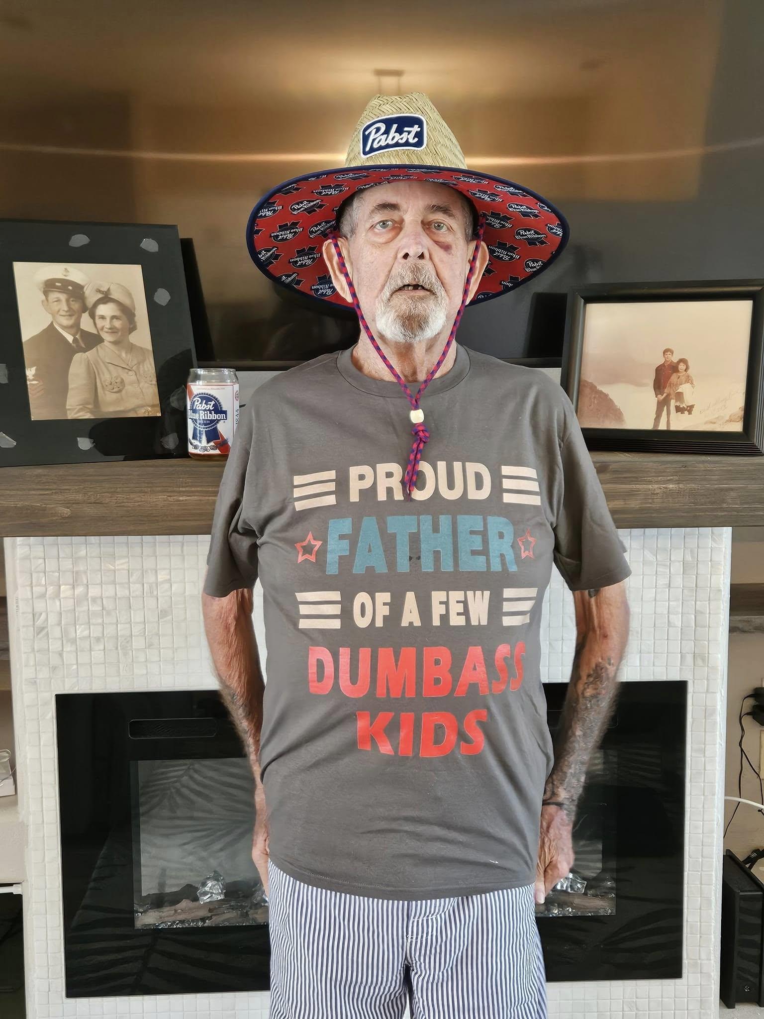 An elderly man poses in a living room wearing a funny shirt and a wide-brimmed hat, smiling.
