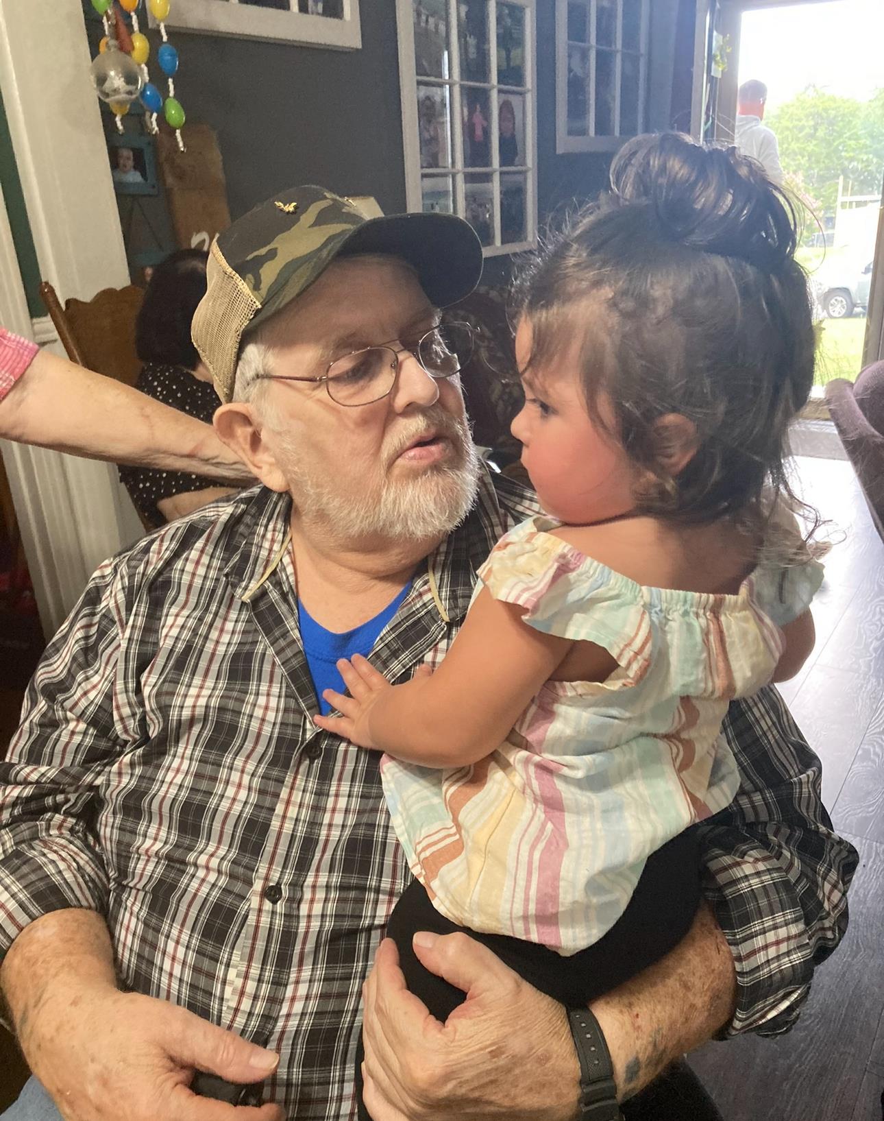 A young girl sits on her grandfather's lap, both smiling and enjoying their time together indoors.