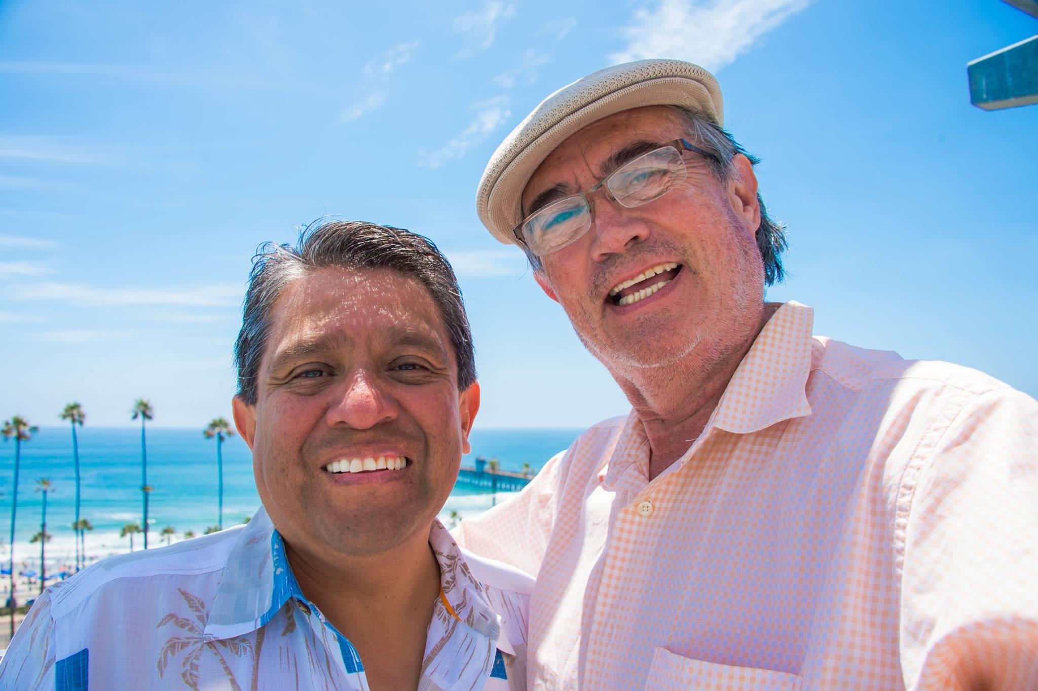 Two men stand close with beaming smiles against the backdrop of a bright ocean and pier.