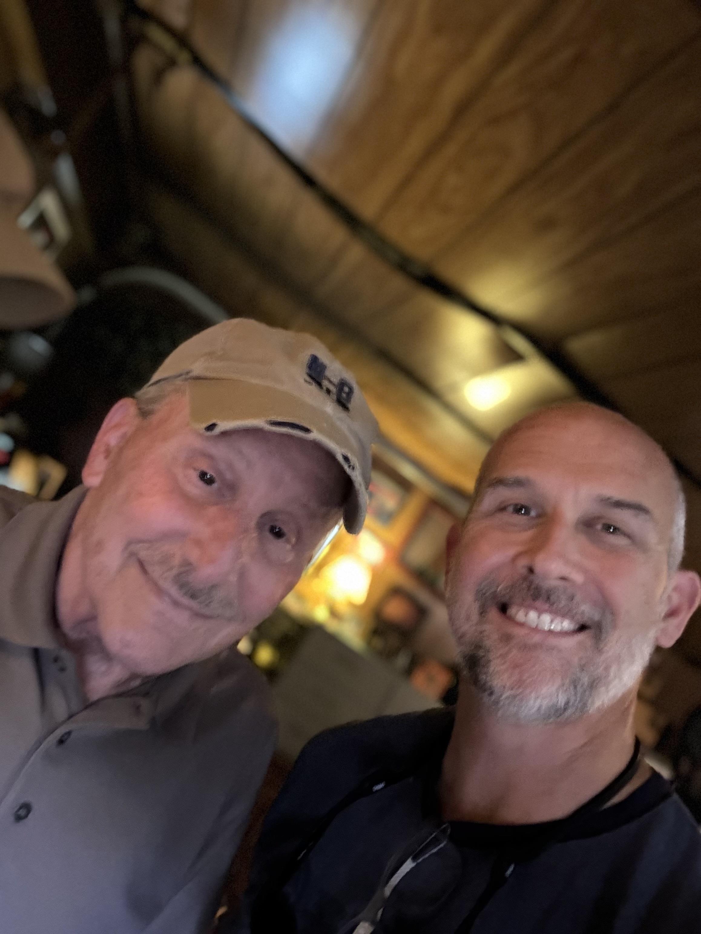 Two smiling men stand close together, sharing a warm moment at a cozy indoor setting.