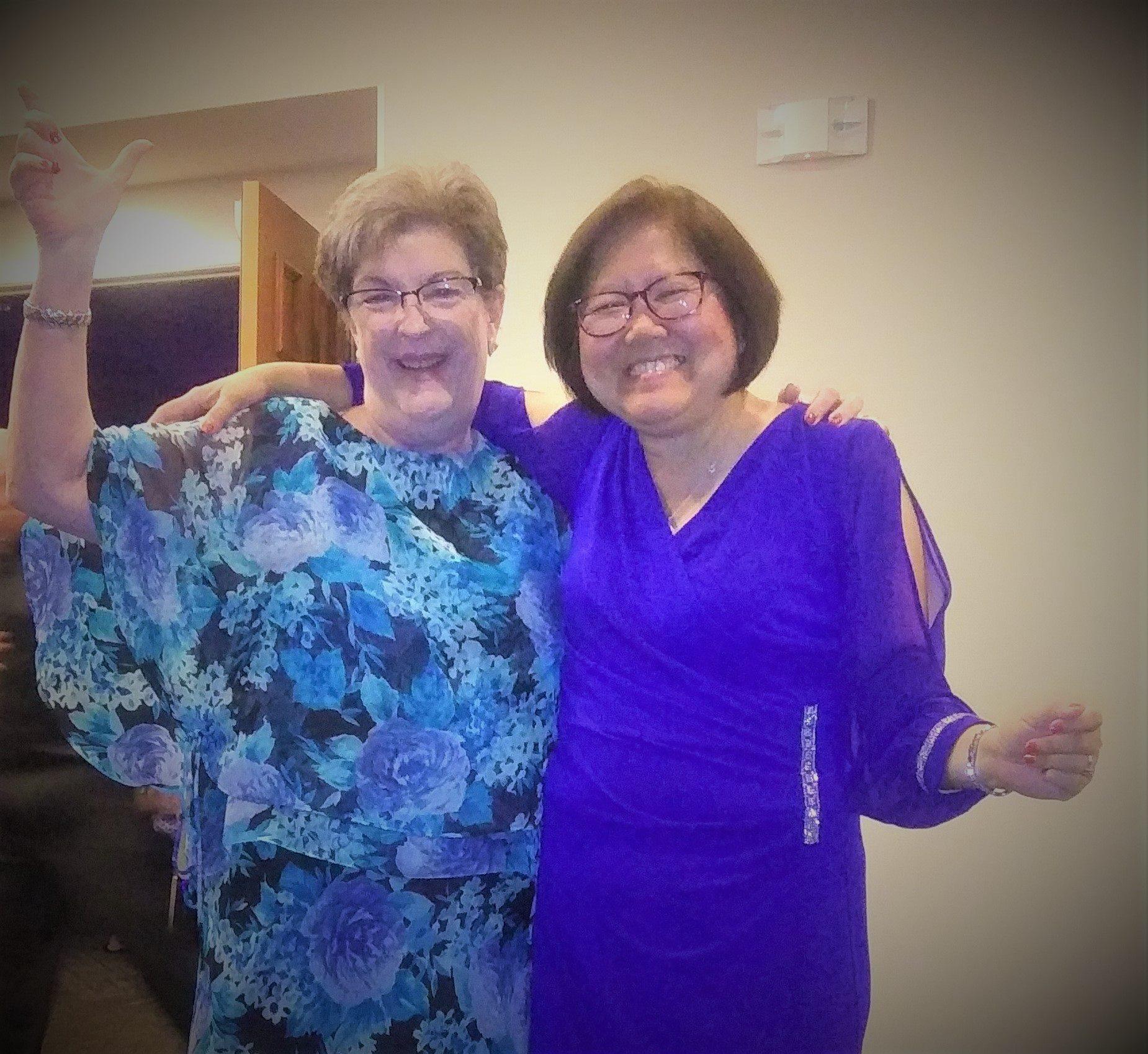 Two women in colorful outfits joyfully laugh together at a warm gathering.