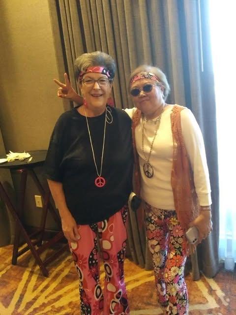 Two elderly women wearing vibrant outfits pose playfully at a festive gathering indoors.