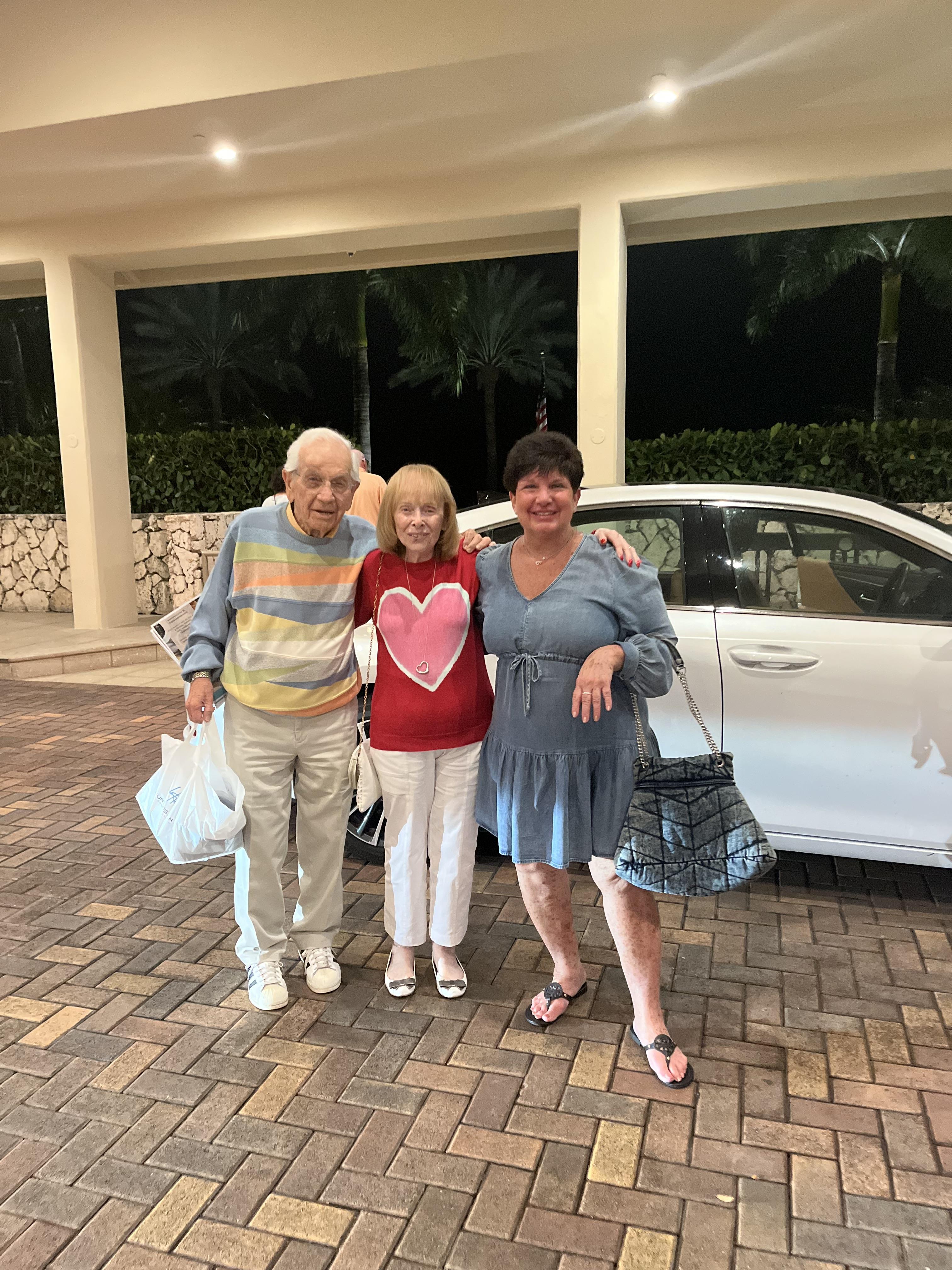 A cheerful elderly couple poses with a friend outside a vehicle after shopping in the evening.