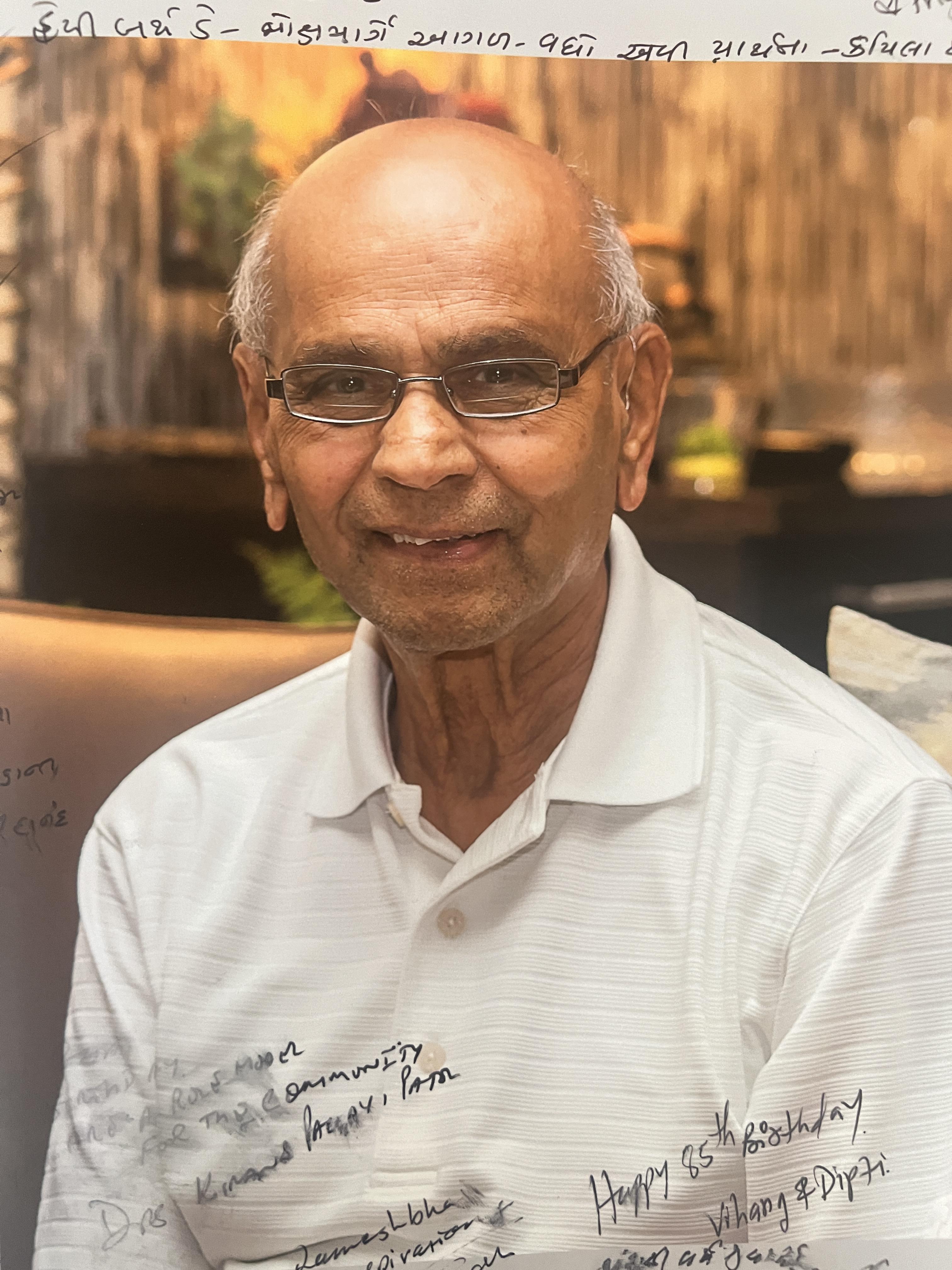 An elderly man with glasses is seated indoors, smiling happily and wearing a white shirt.