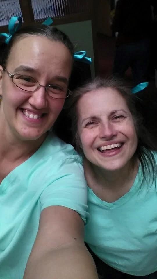Two women in matching teal shirts smile while enjoying a fun moment together.