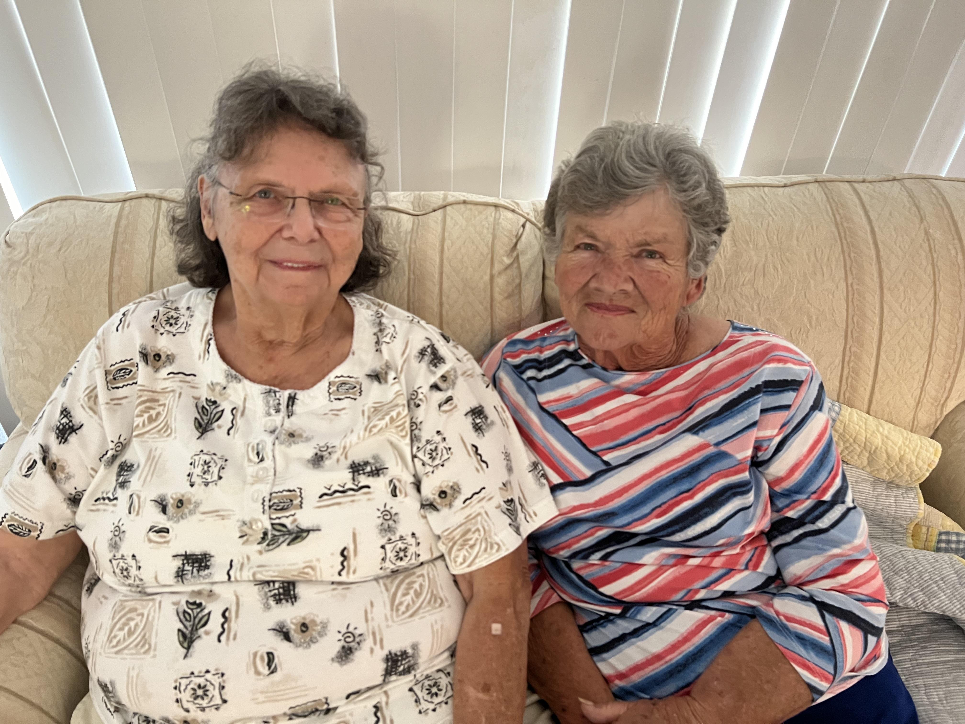 Two elderly women share a cozy moment on a couch, smiling and enjoying each other's company.