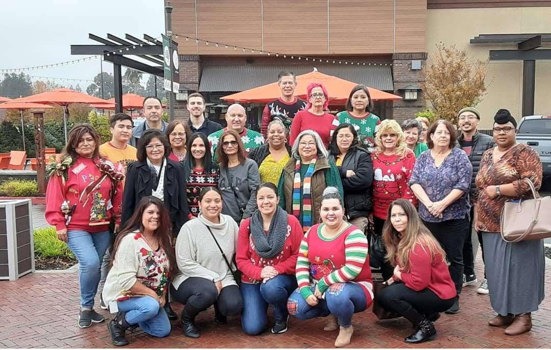Friends gather wearing holiday sweaters at a cheerful outdoor location with festive decorations.