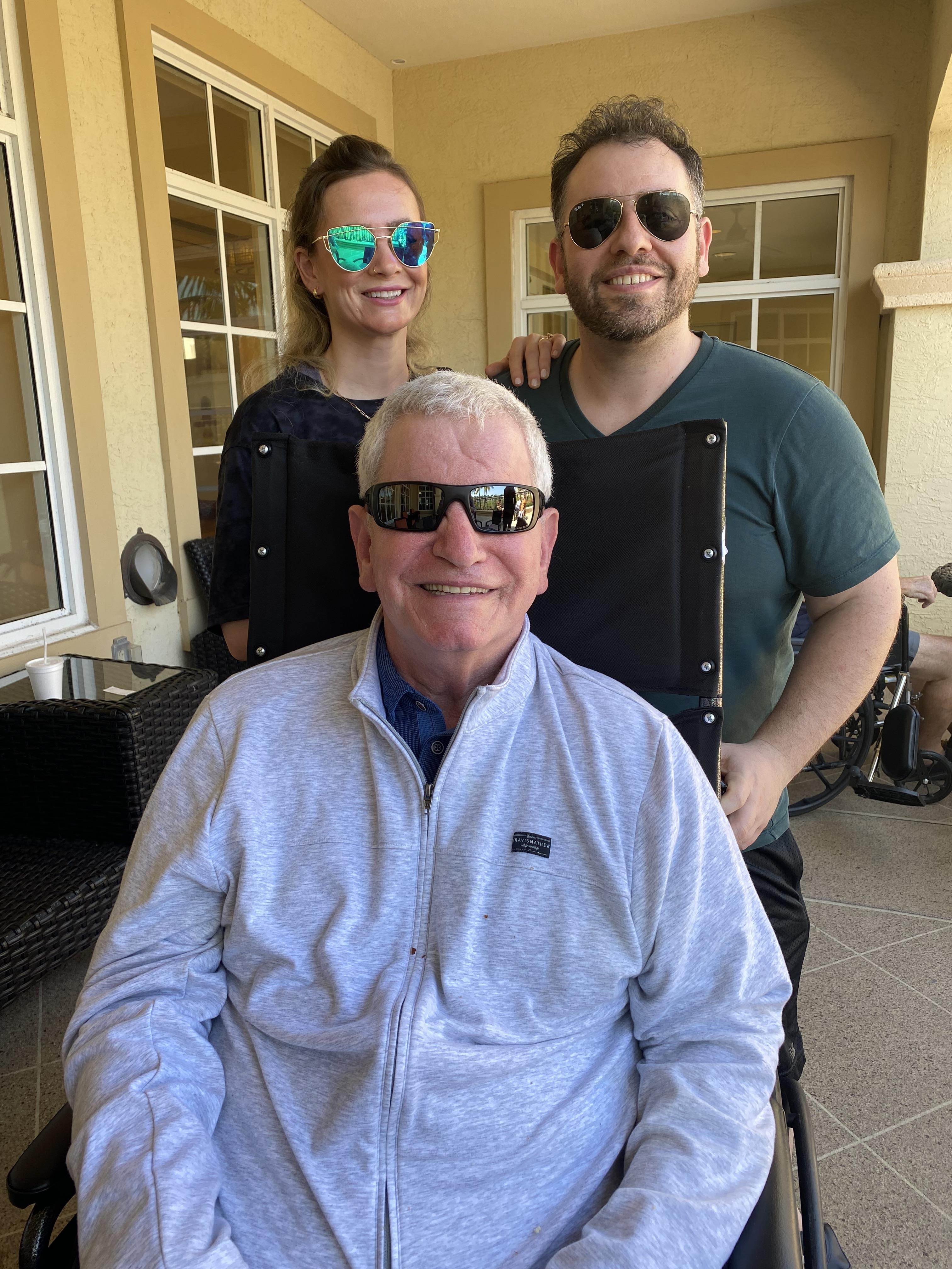 Two adults stand behind a smiling elderly man in a wheelchair, enjoying a sunny day outdoors.