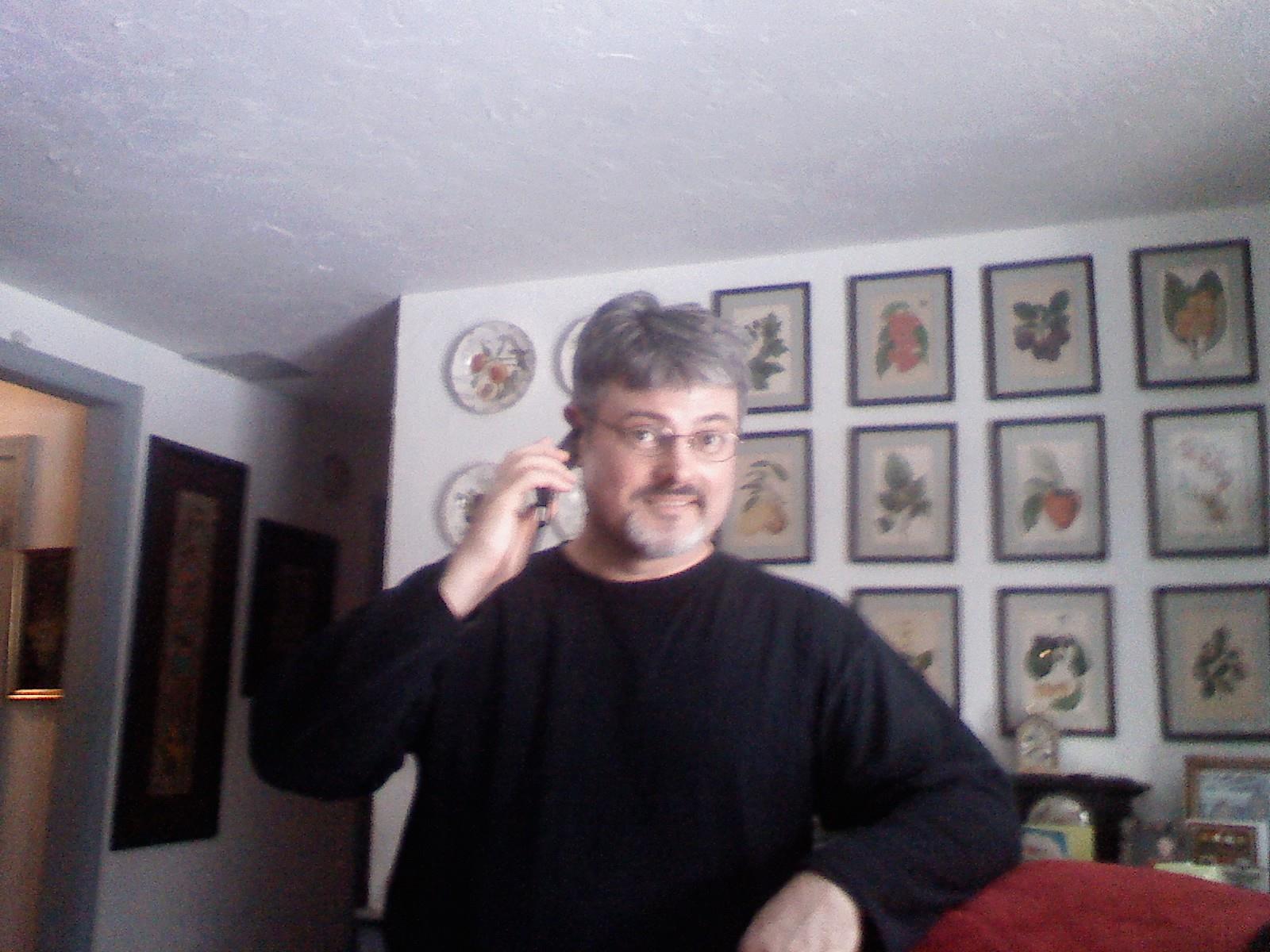 A man is engaged in a phone conversation while standing in a cozy room filled with botanical art.