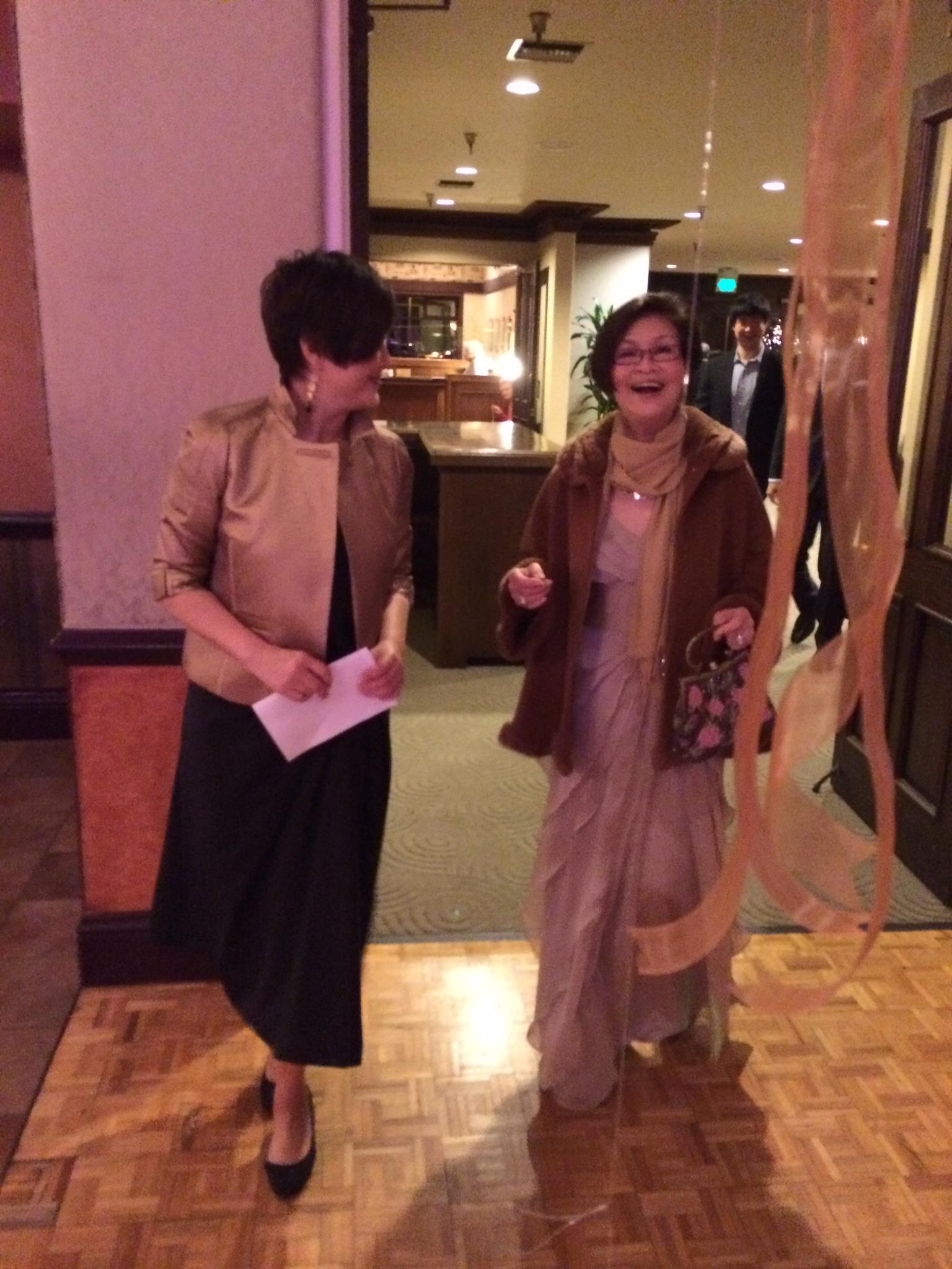 Two women are walking and talking animatedly while dressed elegantly at a festive gathering.