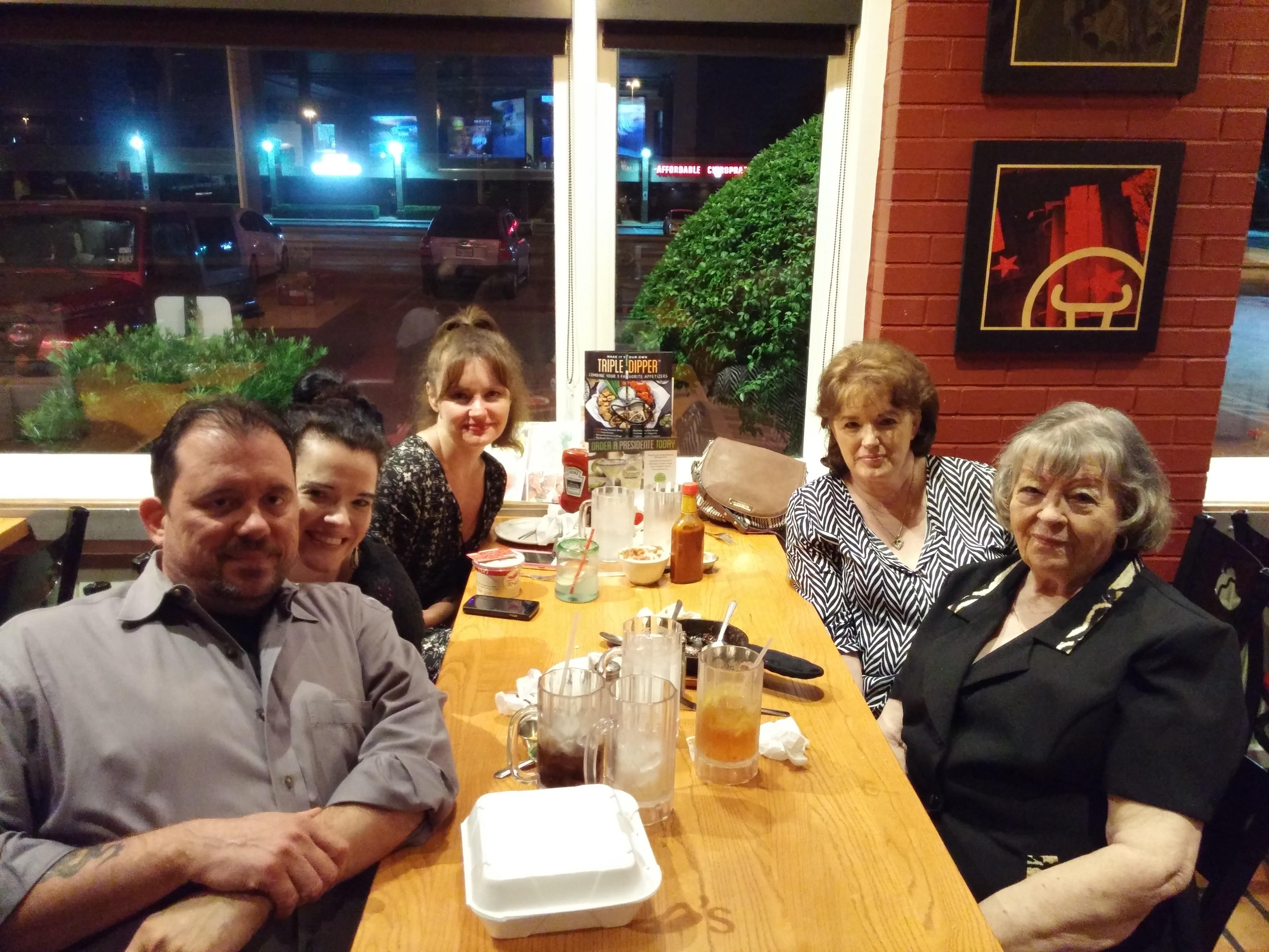 Five people sit around a wooden table, sharing food and drinks while engaged in conversation.
