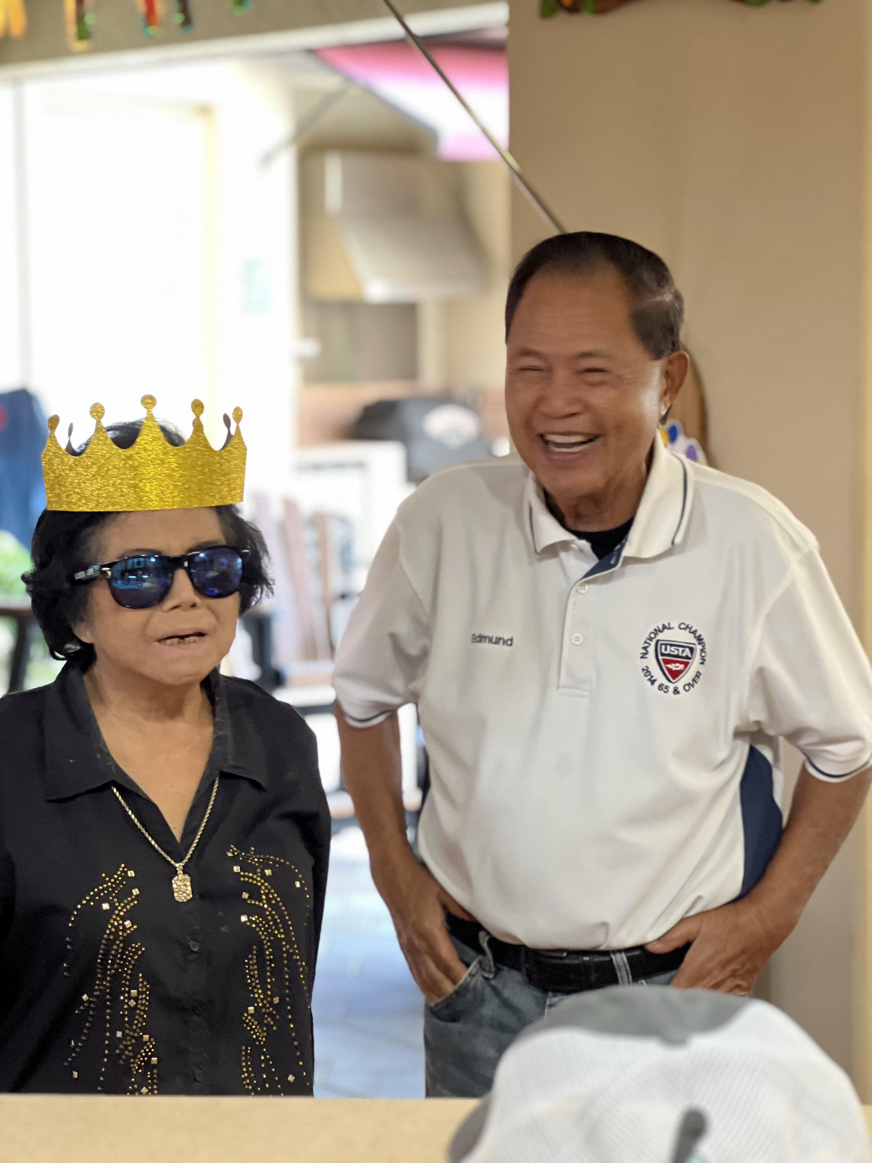 An elderly couple enjoys a light-hearted moment indoors, donning fun accessories while laughing.