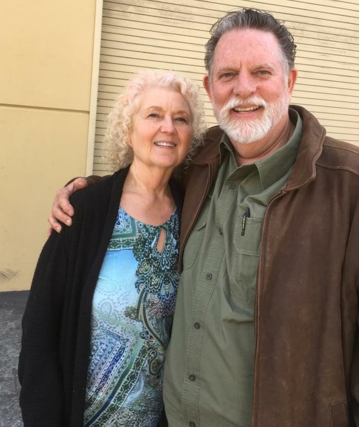 Elderly couple stands closely together, smiling warmly in a casual outdoor environment.
