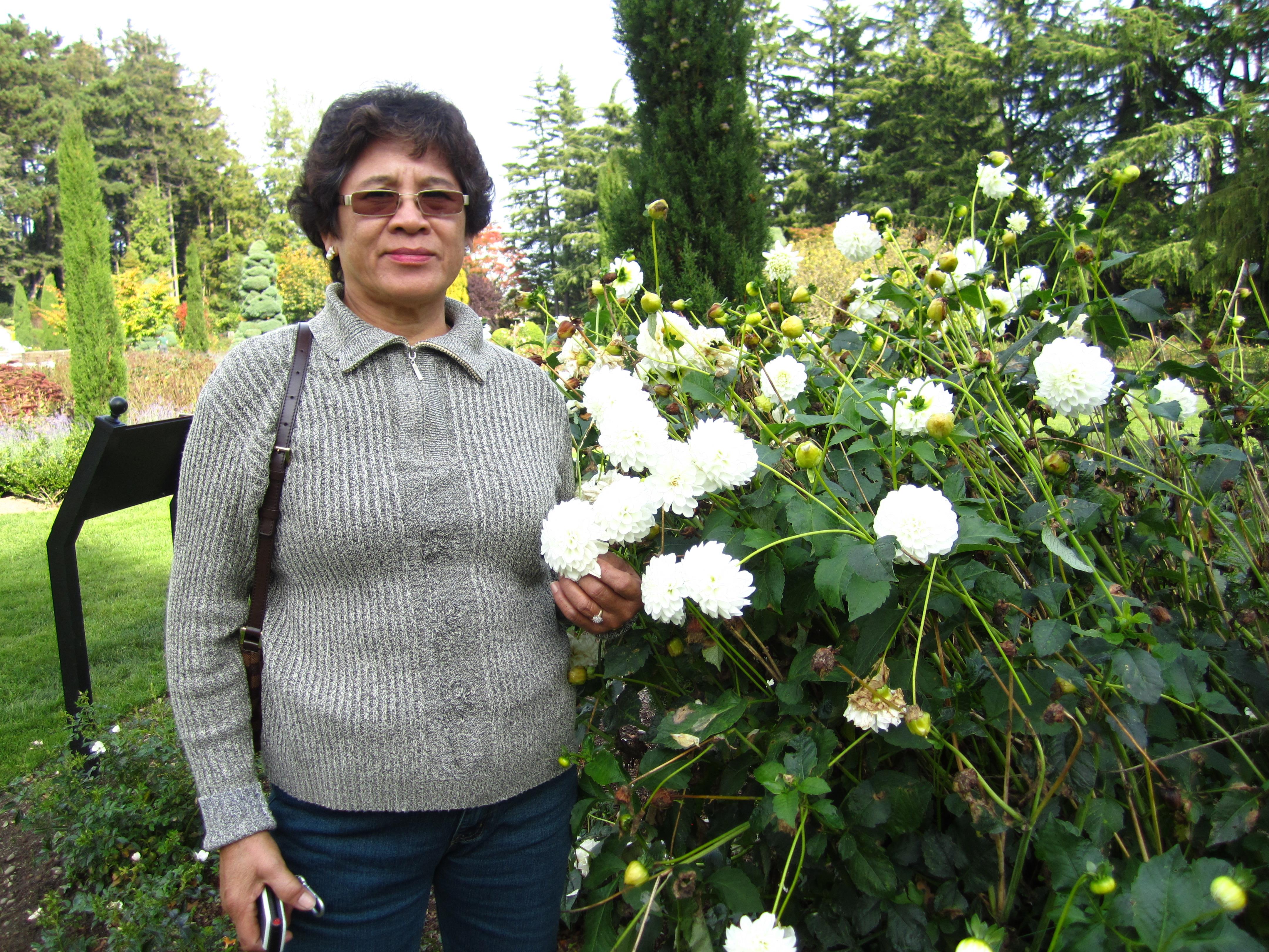 A woman stands next to lush greenery, gently holding a cluster of white flowers while smiling.