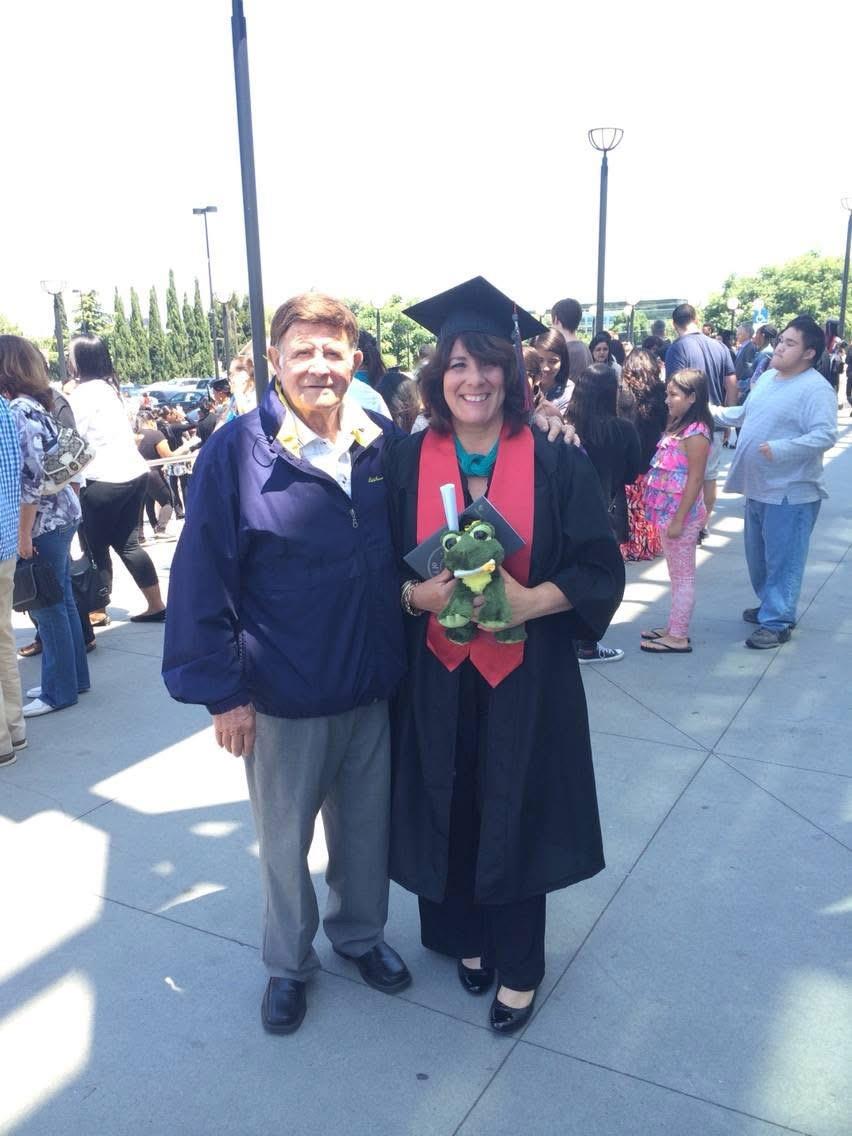 A proud graduate stands with her grandfather, holding a plush toy, amidst a joyful gathering.