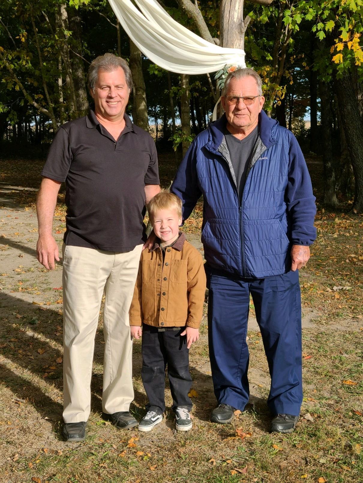 Three generations stand together, enjoying a peaceful moment surrounded by trees and sunlight.