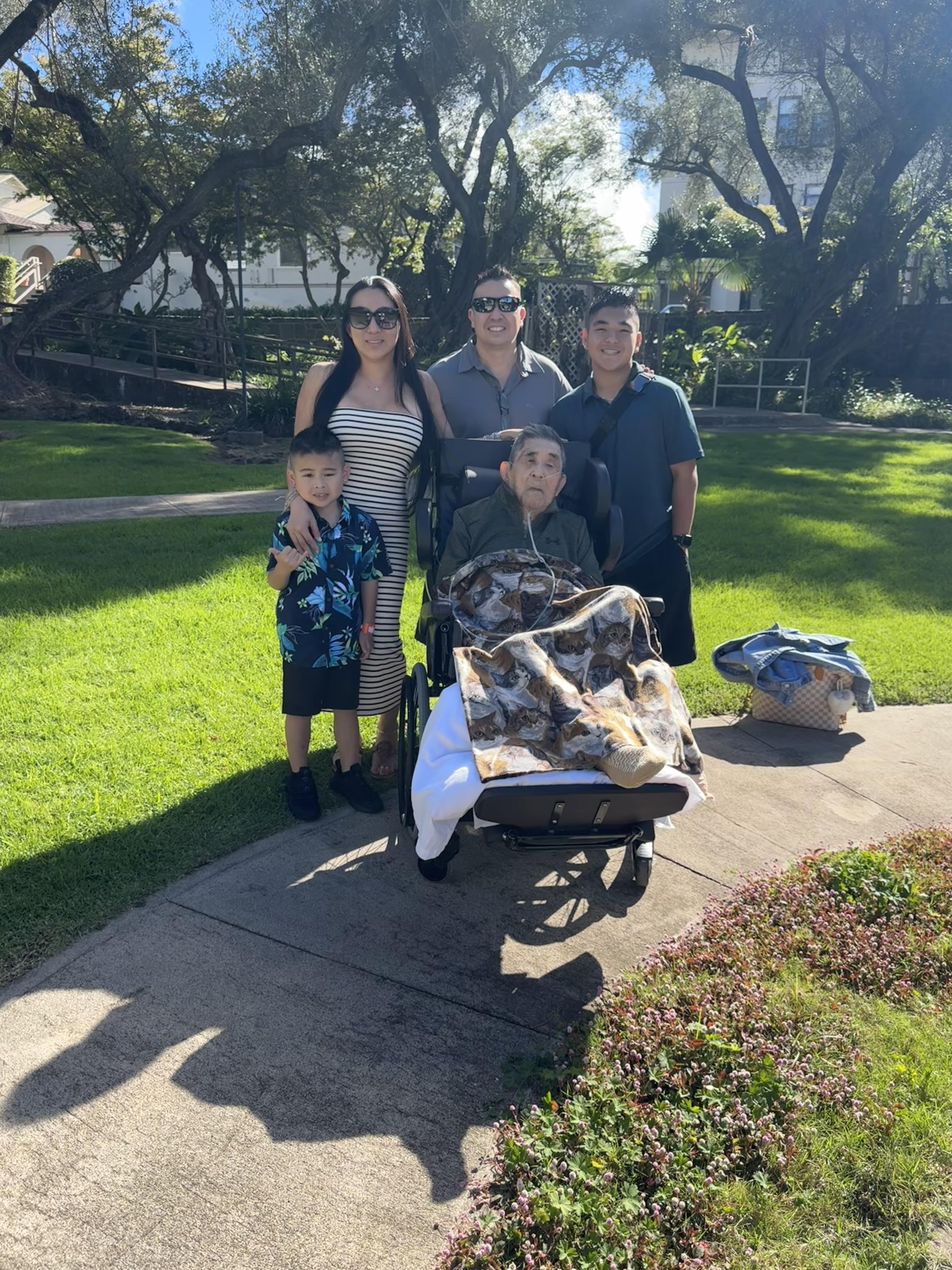 A family enjoys a warm day at the park, smiling together while one member is seated in a wheelchair.