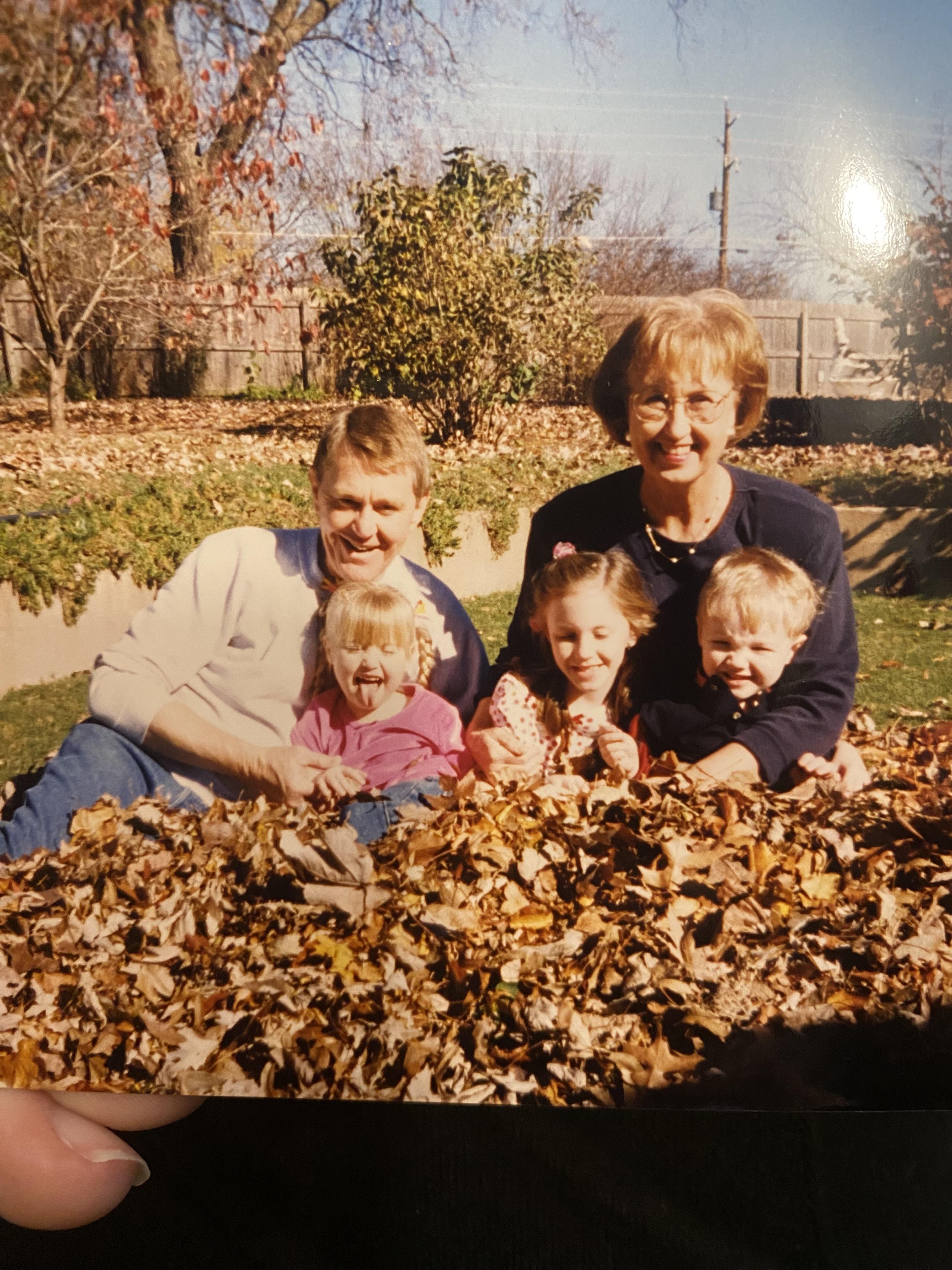 Group of four children and two adults joyfully playing and smiling in a leaf pile on a sunny day.