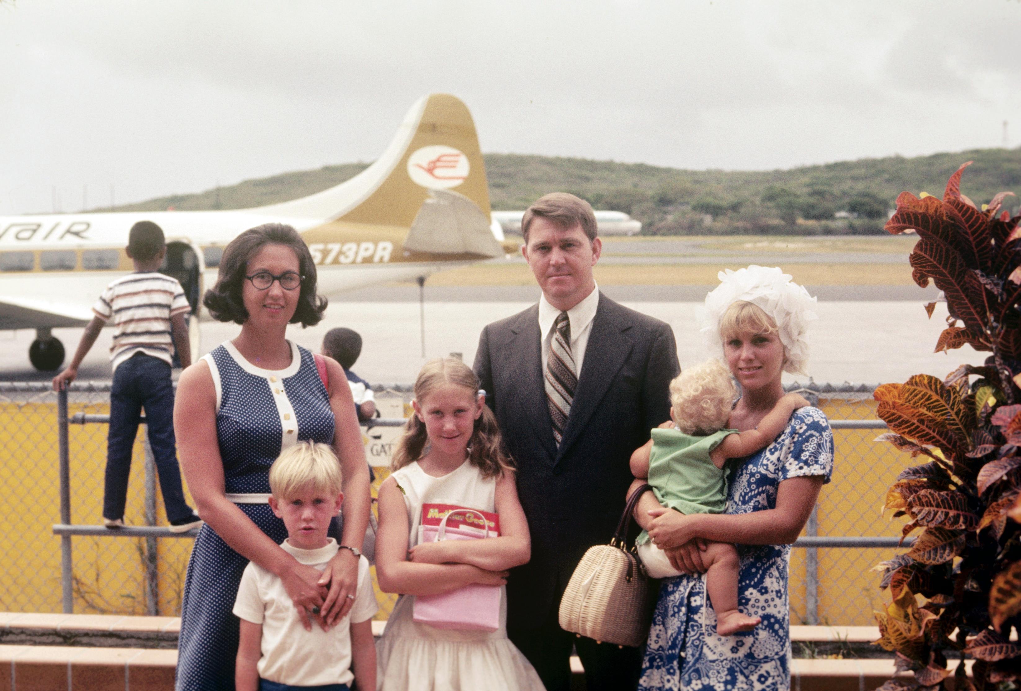 A family poses for a photo at an airport with a vintage airplane behind them on a cloudy day.