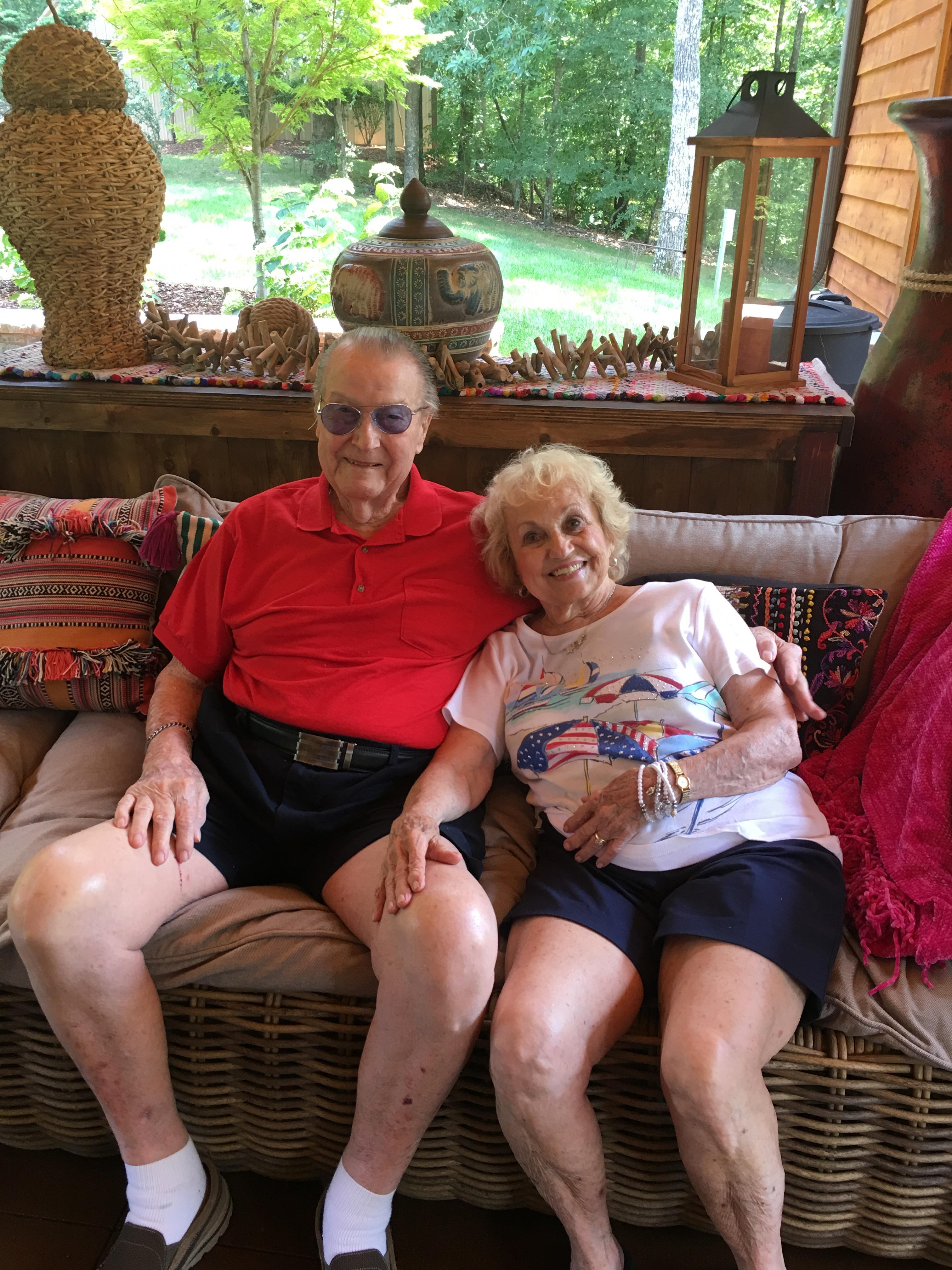 Two seniors sit together on a comfortable couch surrounded by greenery and decor.